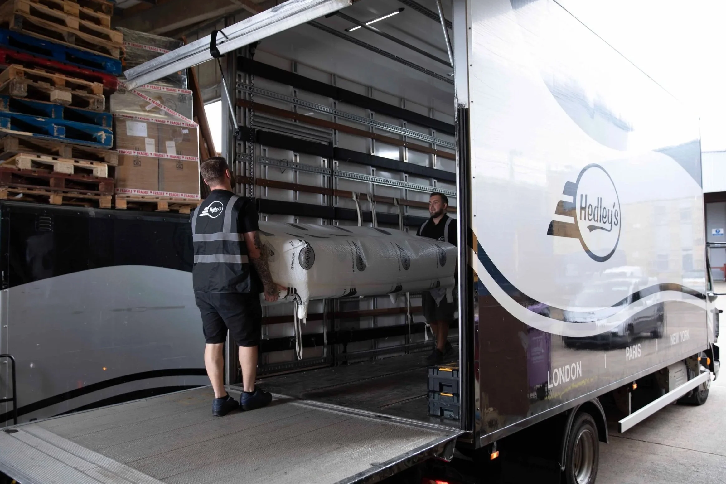 Two men loading a large wrapped pallet into a delivery truck with Hedley's branding, parked near a warehouse with wooden pallets stacked nearby.