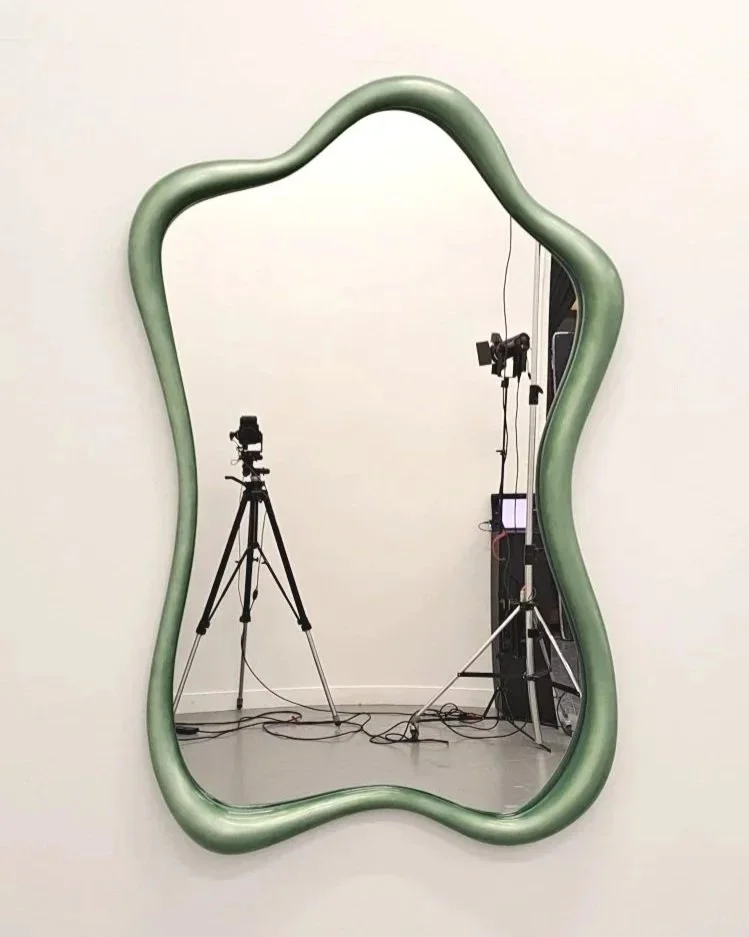 A mirror with an irregular, wavy green frame reflecting a camera setup with tripods and lighting equipment.