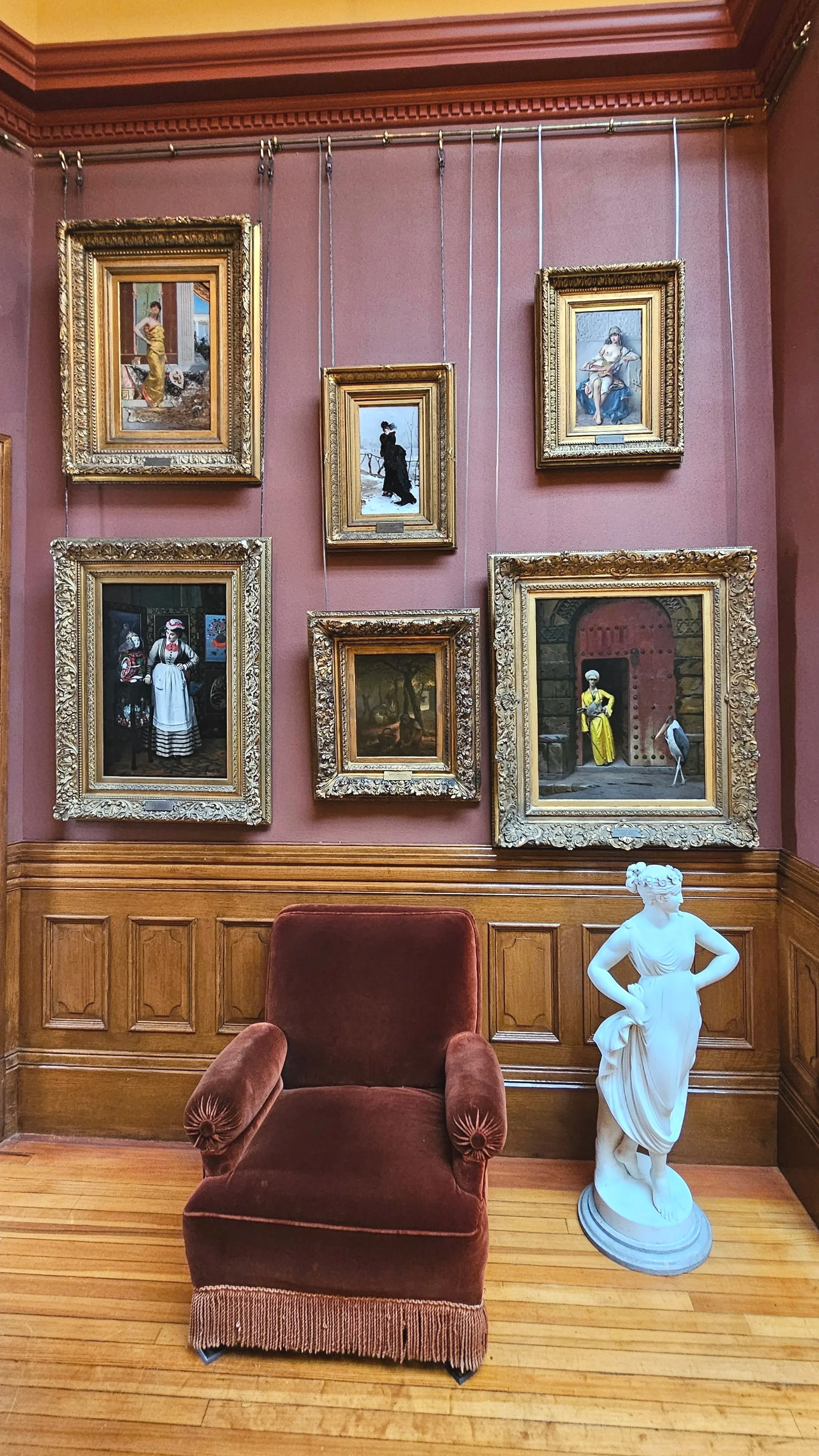 An interior room with pink walls, wooden paneling, and framed art paintings hanging on the wall, a plush red armchair, and a white classical sculpture of a woman.