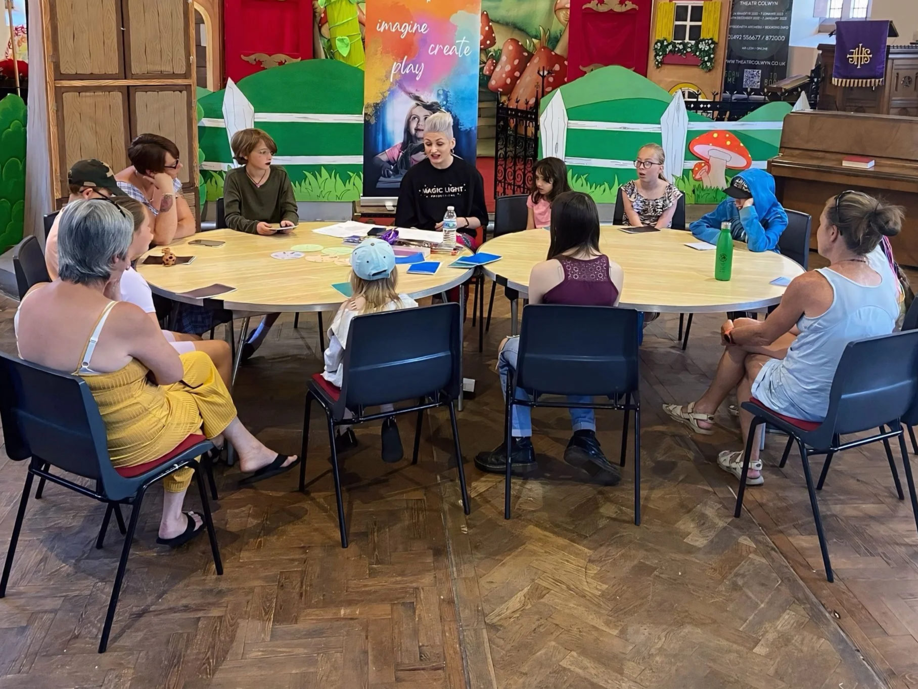 Group of children and adults sitting around a large round table in a colorful, decorated room, attentively listening to a woman with short blonde hair, who is speaking. The room has vibrant artwork and scenery on the walls, and a piano is visible in the background.