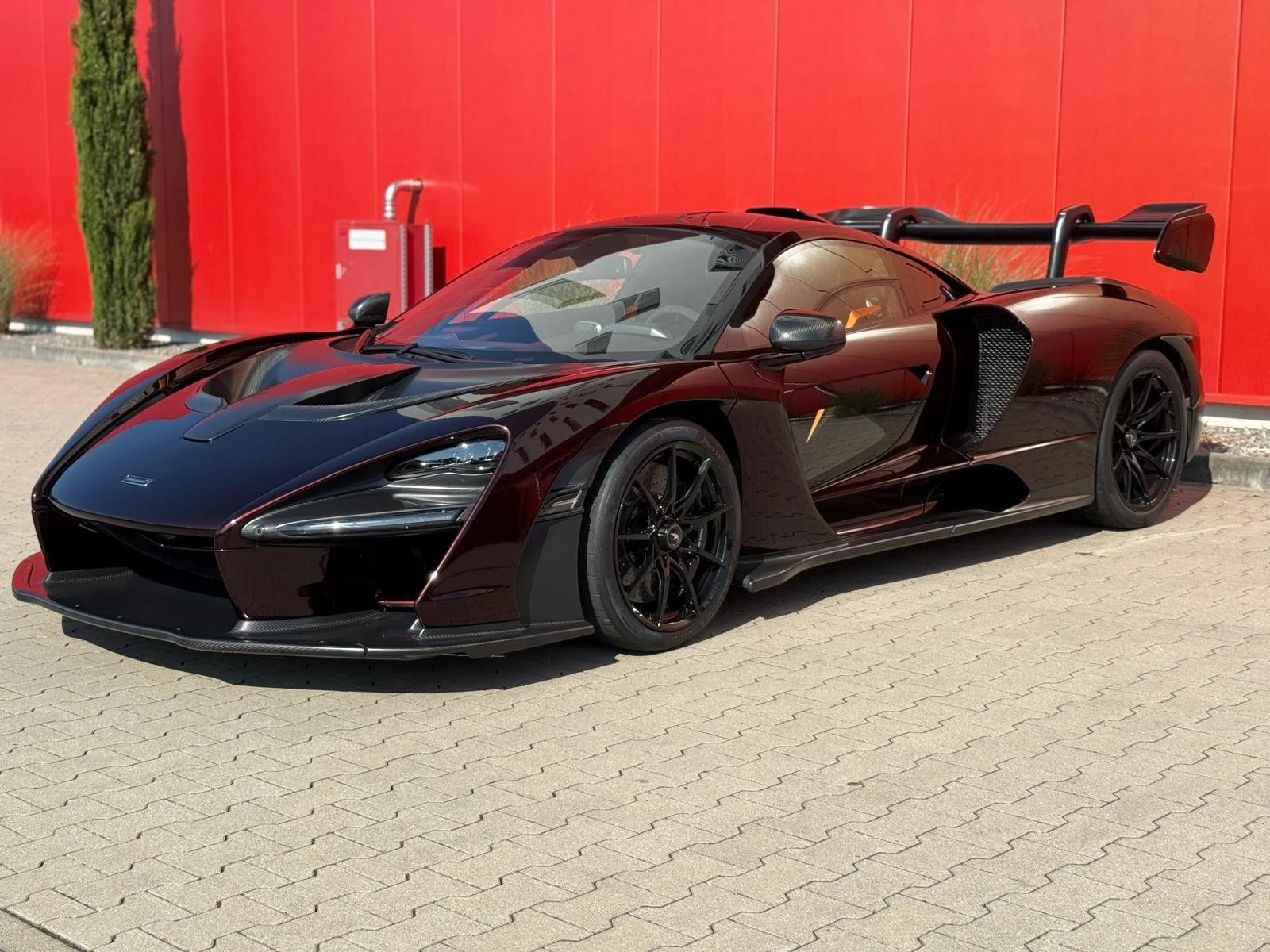 Une voiture de course noire avec des détails rouges, stationnée devant un mur rouge, avec un grand aileron arrière.