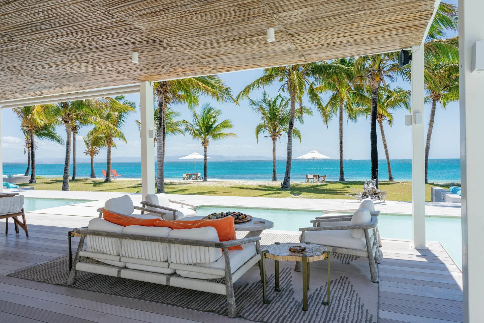 Beachside patio with white seating, orange and white cushions, overlooking palm trees, ocean, and blue sky with umbrellas in the distance.