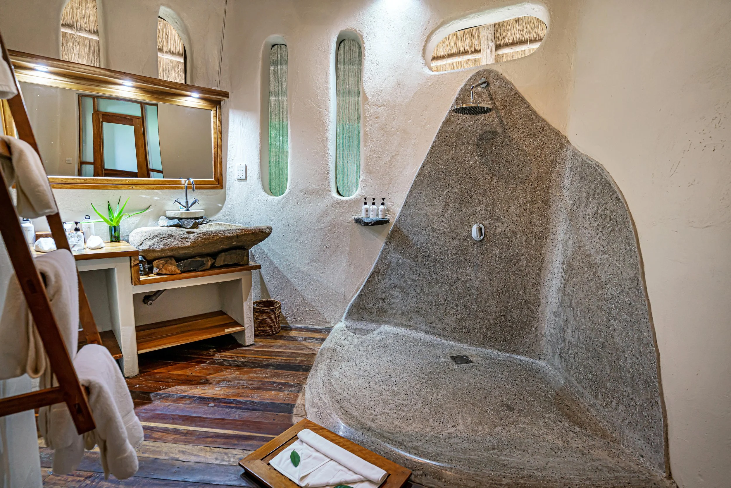 Bathroom with a large rounded shower with a rain showerhead, a stone sink with a faucet, a mirror, wooden shelves, towels, and small bottles of toiletries, with wooden floors and windows with textured glass.