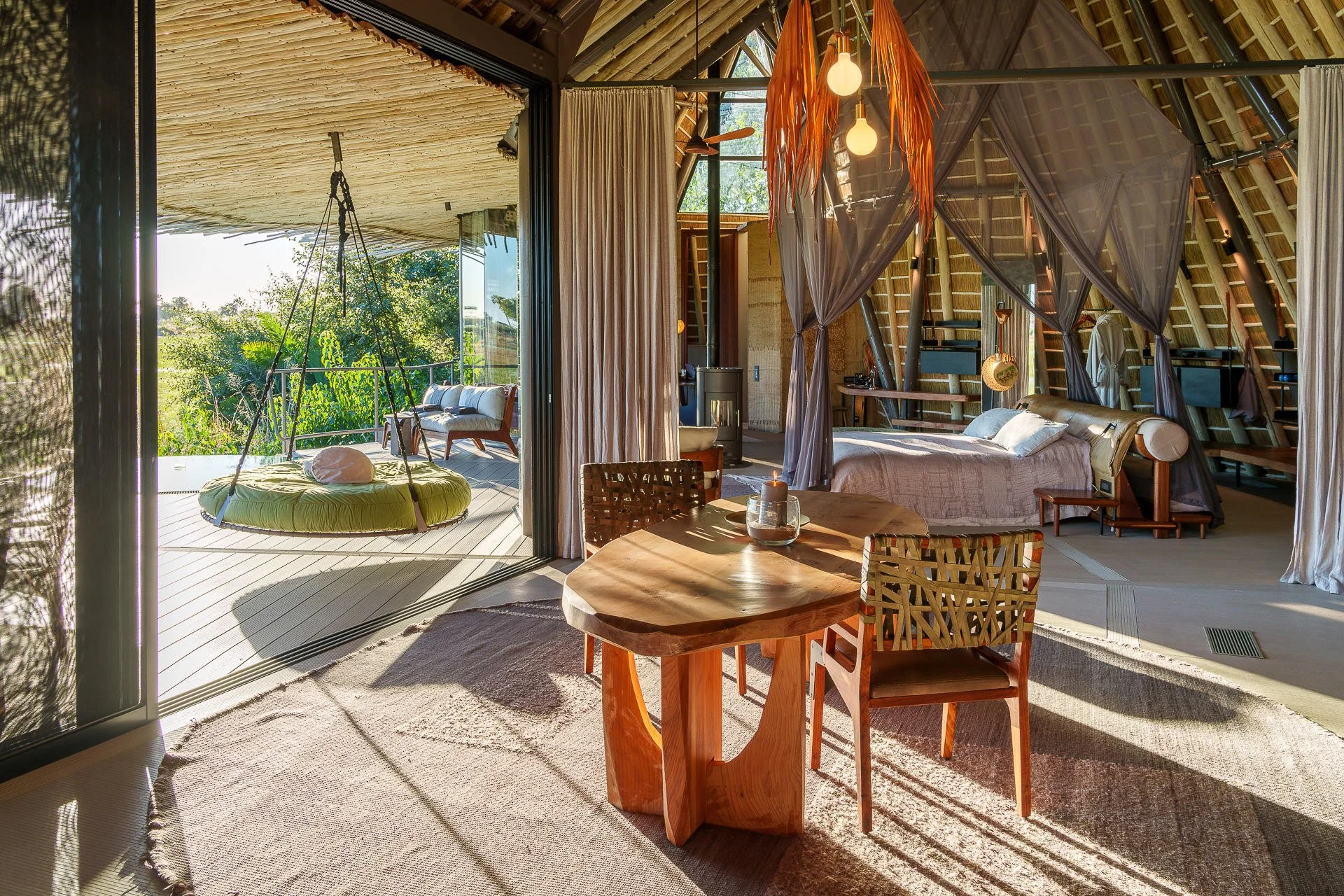 Interior view of a cozy, rustic bedroom with a wooden canopy bed covered in neutral bedding, draped curtains, a sitting area with outdoor furniture on the balcony, and warm lighting including hanging bulbs and a pendant lamp.