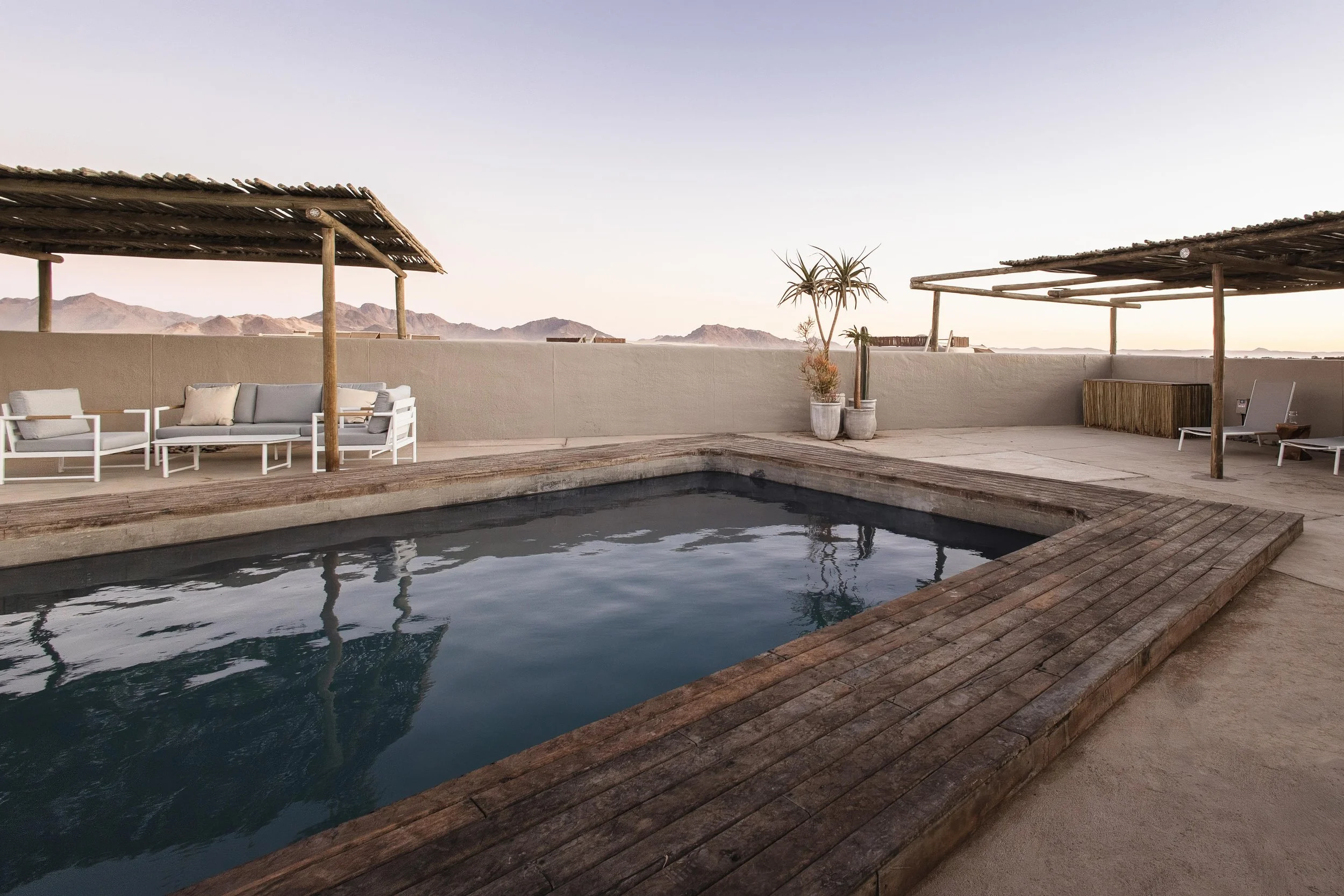 A rooftop pool area with lounge chairs, sun umbrellas, and potted plants, overlooking a desert landscape with mountains in the distance during sunset.