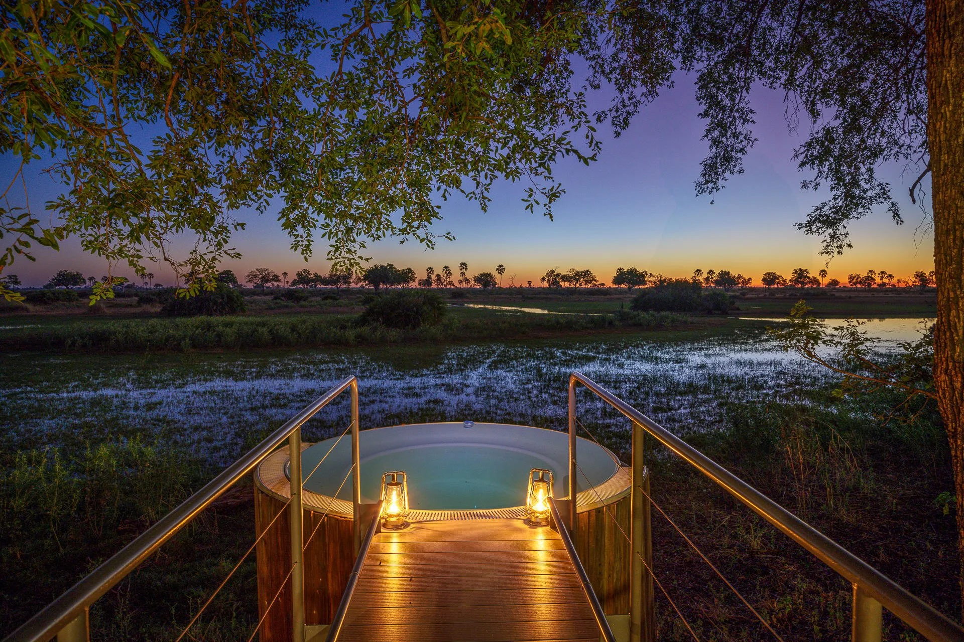 Outdoor hot tub at dusk with lanterns on a wooden deck, overlooking a river and a sunset sky, surrounded by trees.