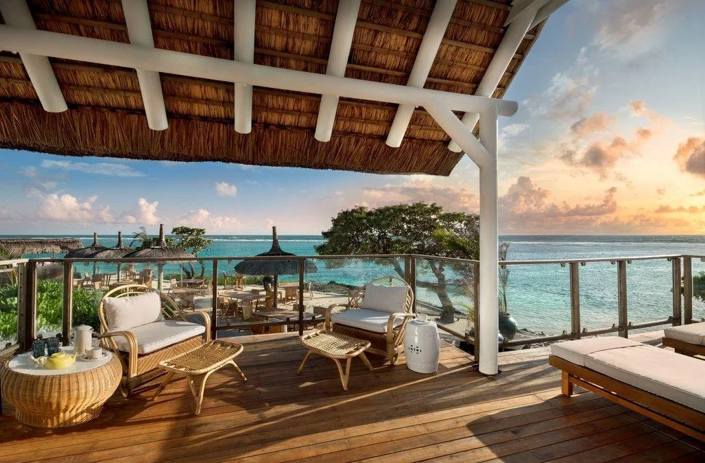 Beachside resort balcony with wicker furniture, lounge chairs, and umbrellas overlooking the ocean during sunset.