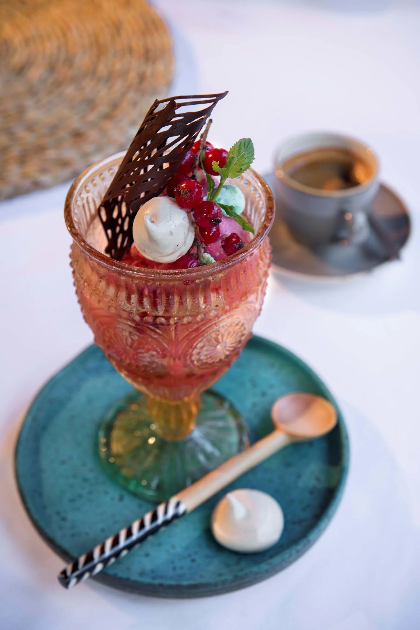 A glass dish with pink dessert topped with red berries, a mint leaf, and a chocolate garnish, served on a teal plate with a small spoon and dollop of cream, with a cup of coffee in the background.