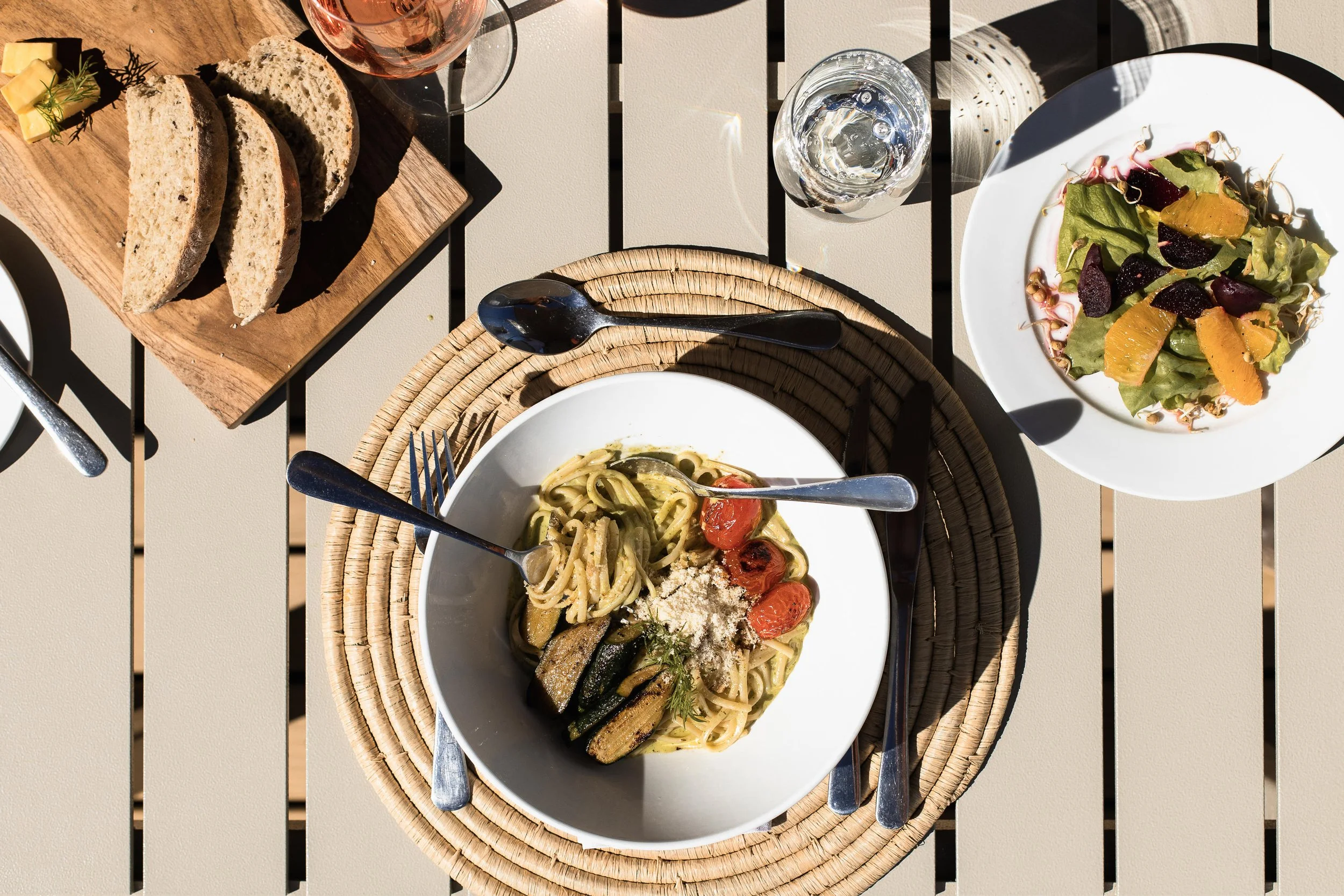 A top-down view of a meal on a beige slatted outdoor table, featuring a bowl of pasta with zucchini, cherry tomatoes, grated cheese, and herbs, a side salad with greens, orange slices, and beets, a wooden board with slices of bread, a glass of water,