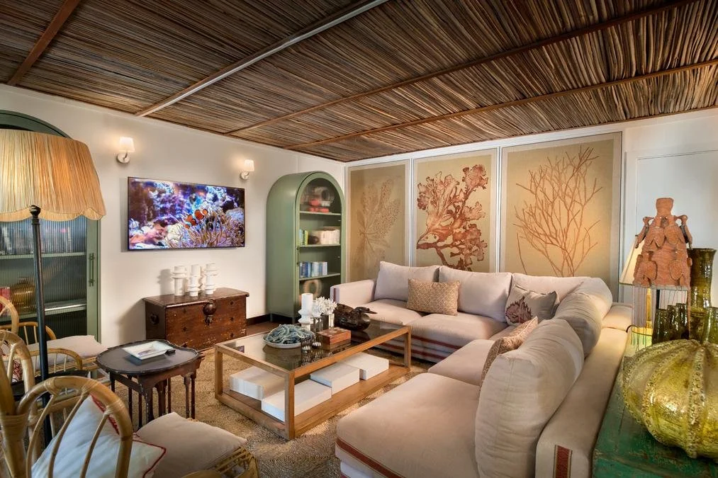 Living room with beige sectional sofa, wooden coffee table, green bookshelf, wall art of coral illustrations, wooden ceiling, and various decorative items.