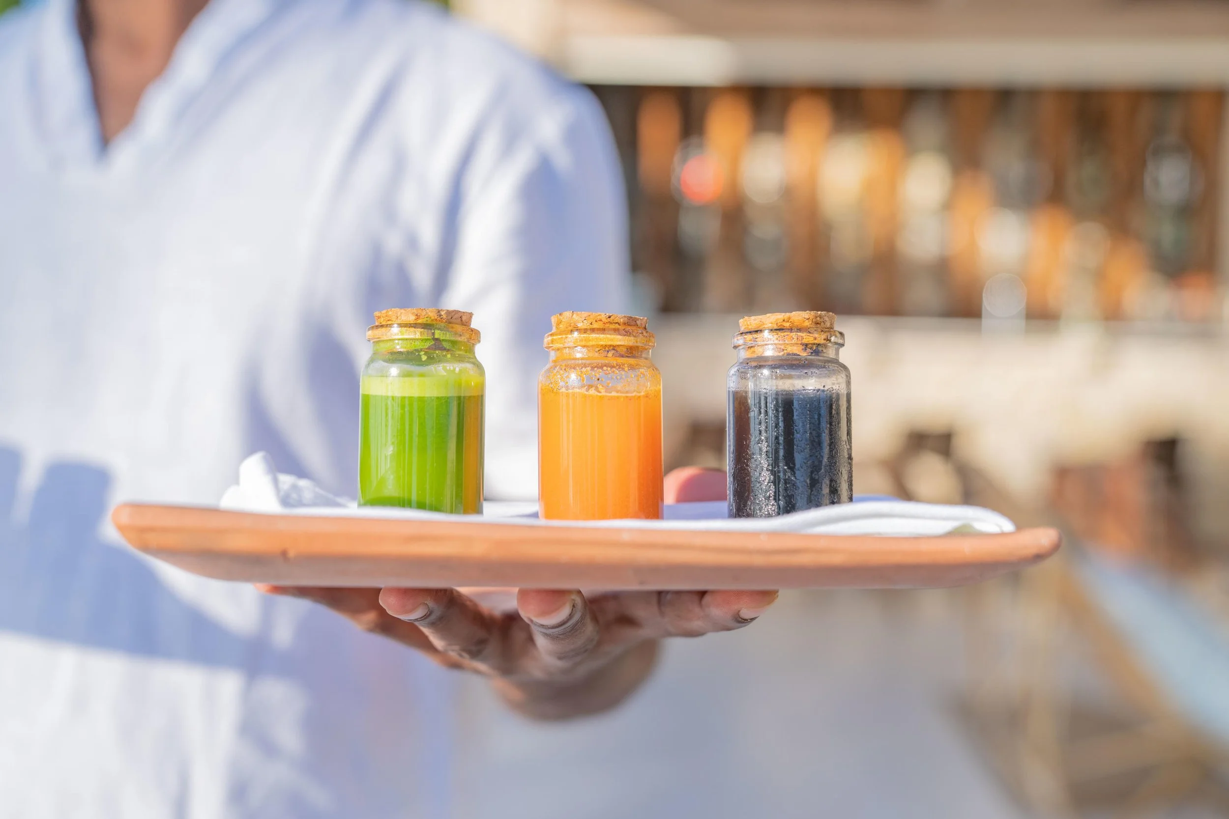 A person holding a wooden tray with three small glass bottles containing colored liquids: green, orange, and black, with cork stoppers, outdoors.