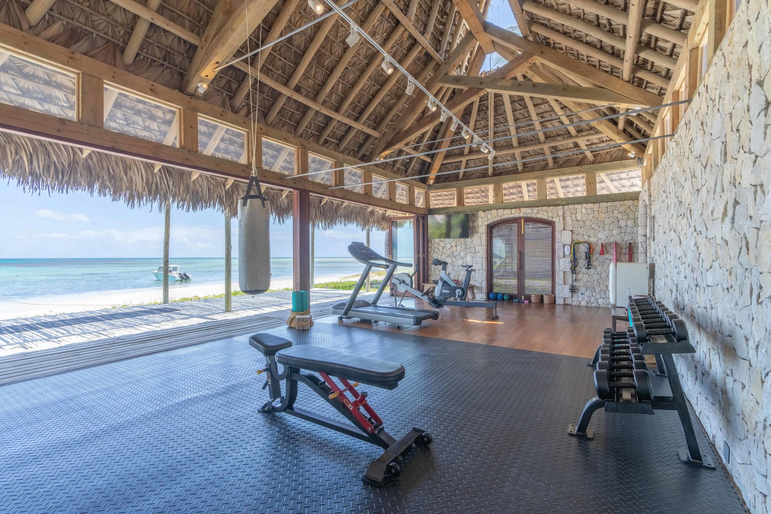 Beachside gym with exercise equipment including a treadmill, jump rope, dumbbells, and a punching bag, overlooking the beach with a boat on the water.