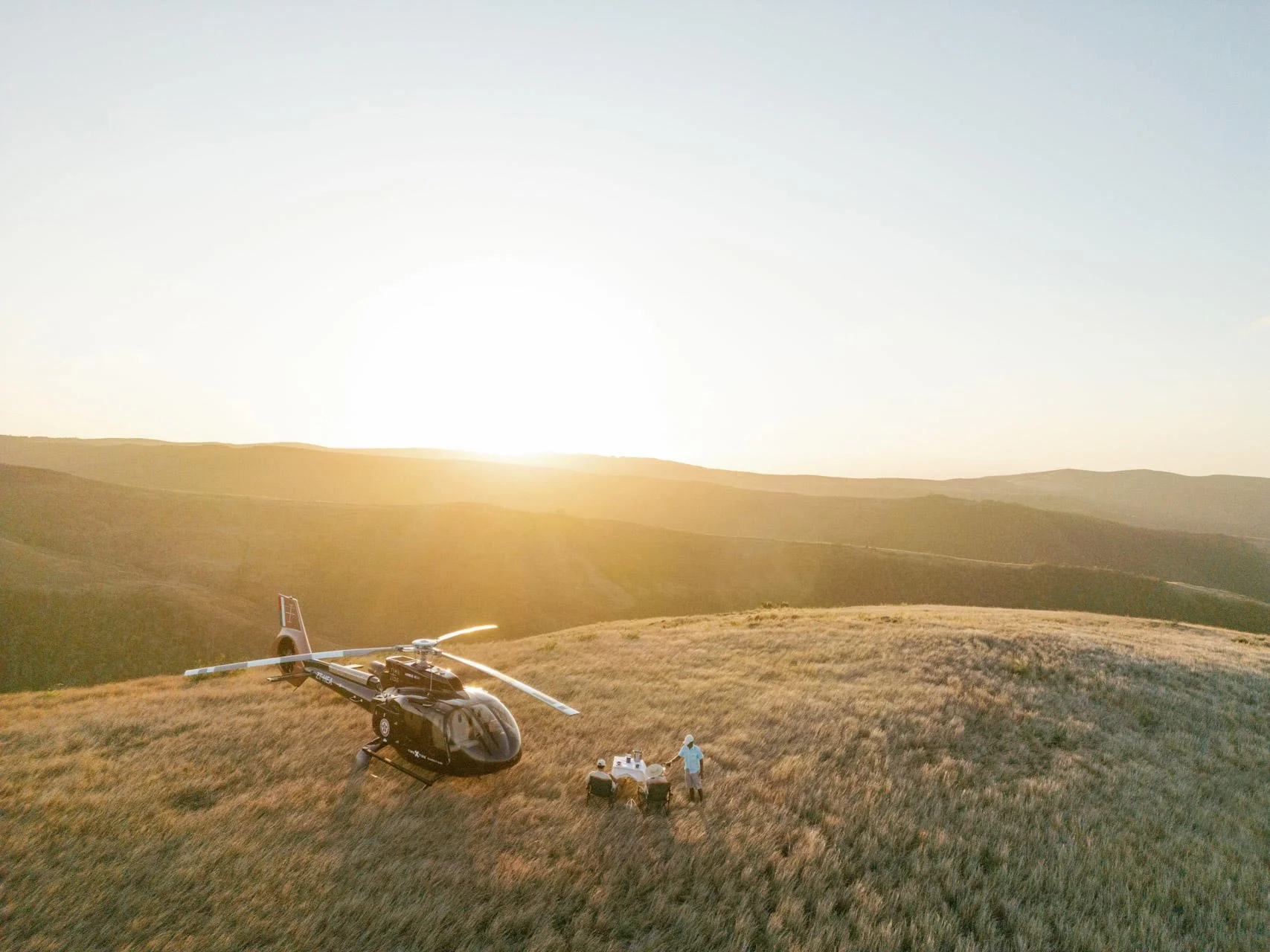 A black helicopter landing on a grassy hill during sunset, with two people having a meal at a small table nearby.