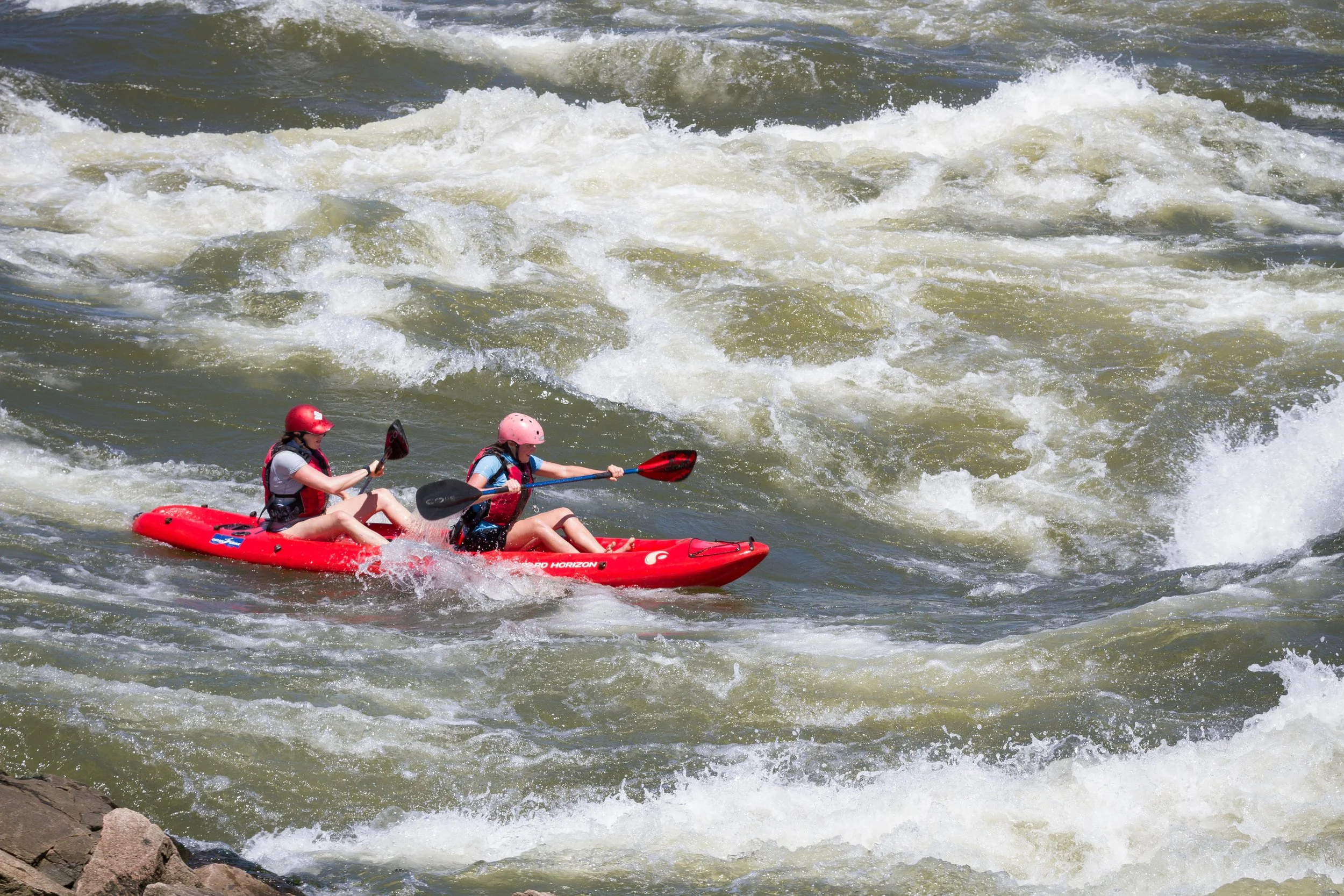 Two people kayaking in rough water with waves and rocks in the foreground.
