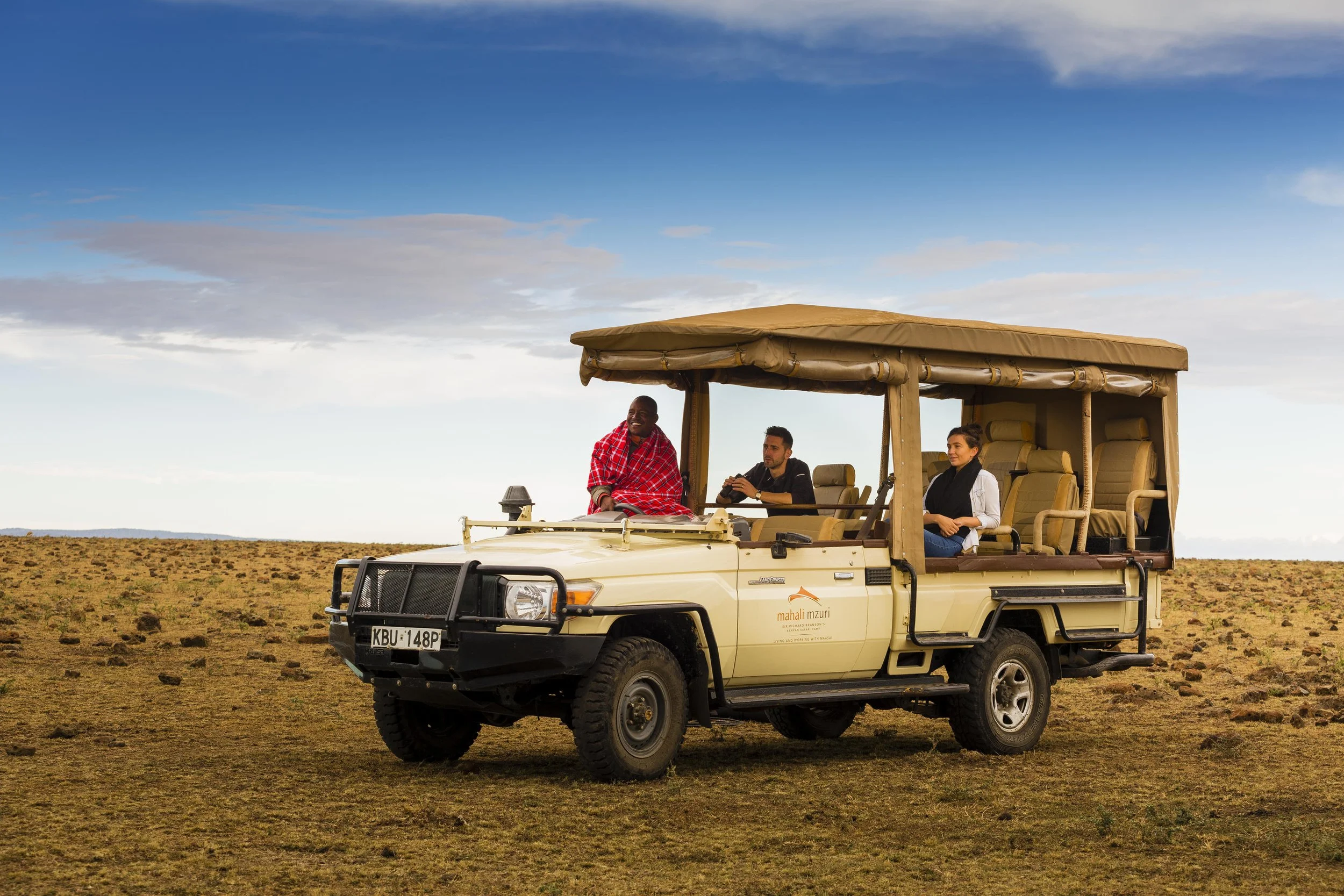 Three people riding in an open safari vehicle on a grassy plain during daytime.