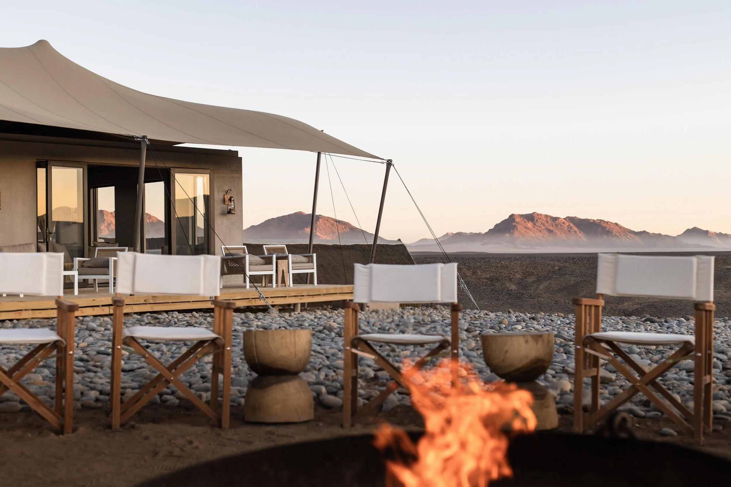 Outdoor scenic view of a modern desert house with patio furniture, large shade sail, and fire pit, set against rocky mountains at sunset.
