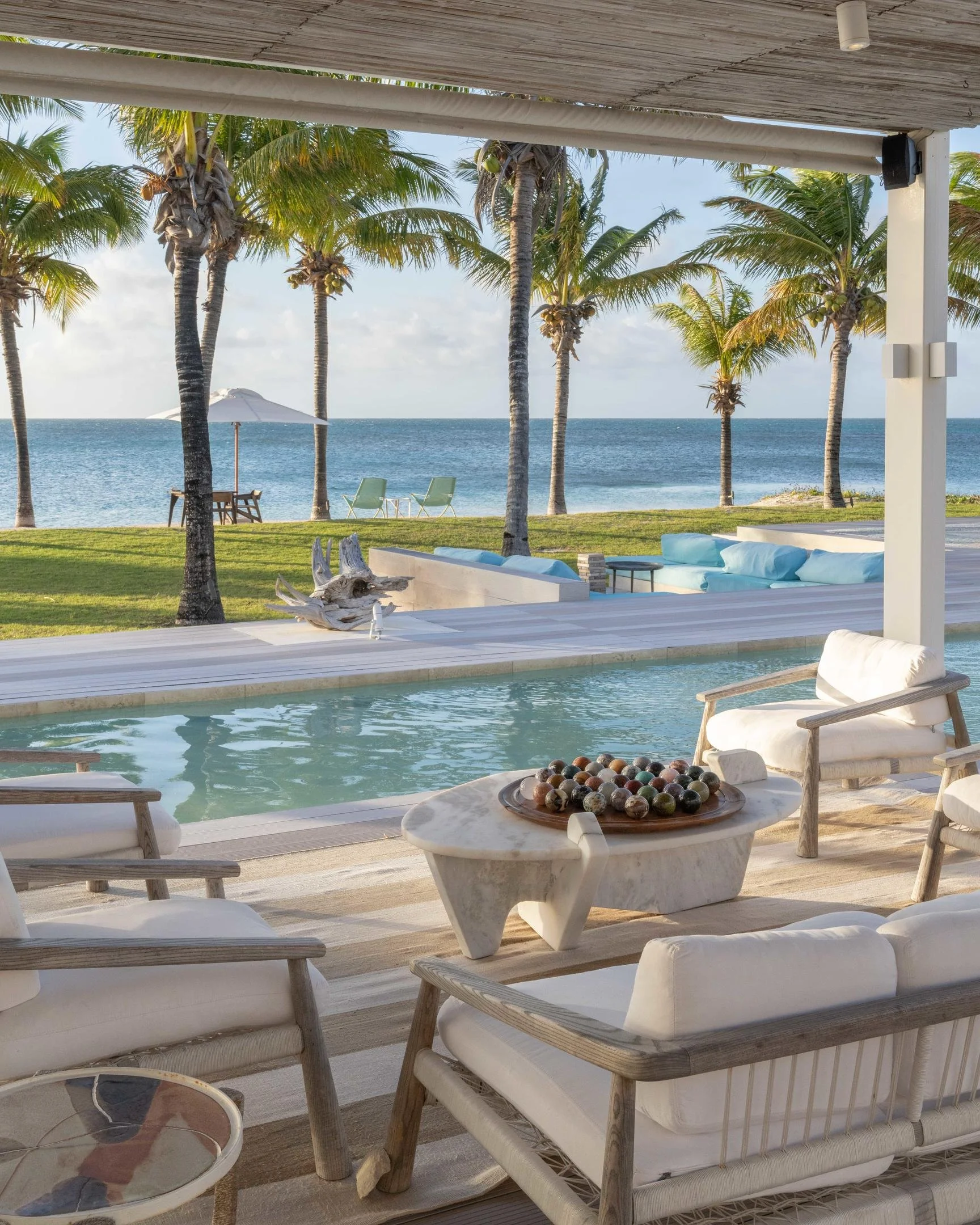 A luxurious outdoor seating area overlooking a pool and the ocean, with palm trees, lounge chairs, and an umbrella in the background.