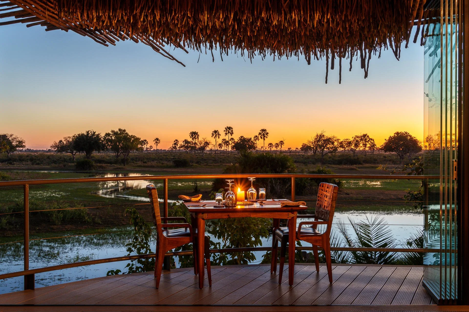 A dining table set for dinner on a balcony overlooking a river and sunset with trees in the background.