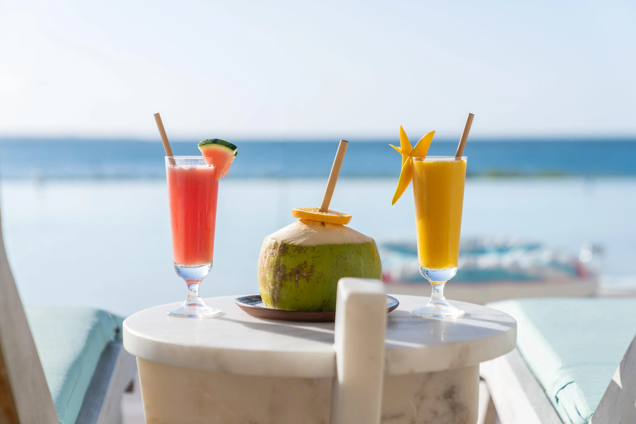 Two tropical fruit drinks and a fresh coconut with a straw on a table by the ocean, with boat in the background