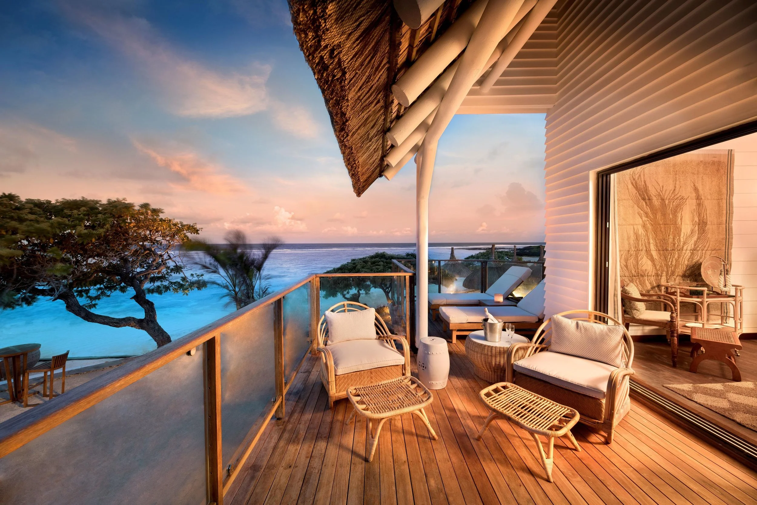 A cozy outdoor balcony with wooden flooring and rattan furniture overlooking the ocean at sunset, with trees and pink clouds in the background.
