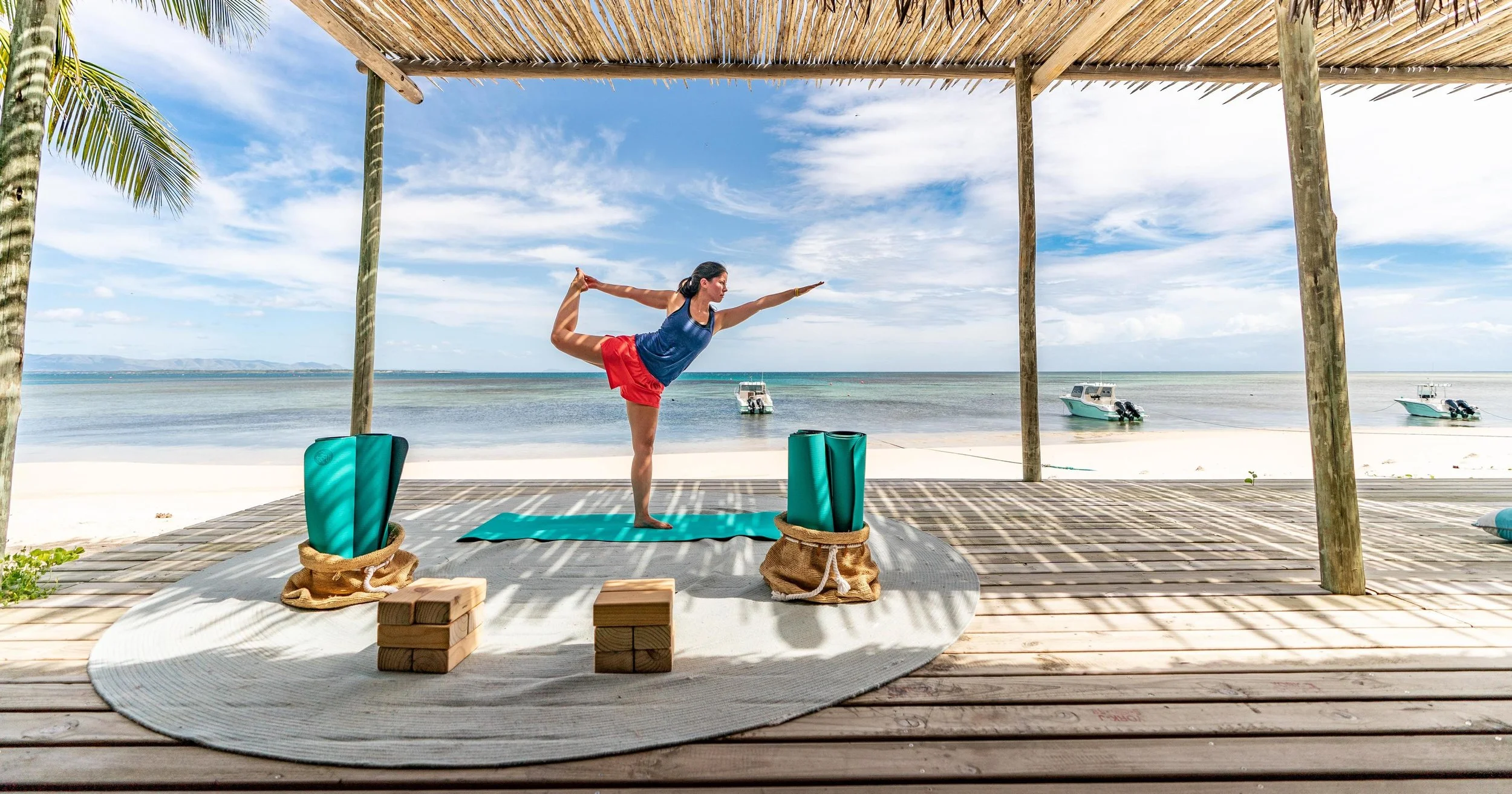 A woman practicing yoga on a mat on a beach covered by a wooden and thatch canopy with boats in the water in the background.