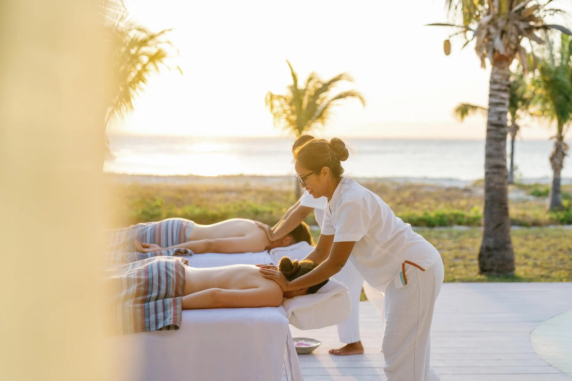 Two people receiving massage outdoors near the beach at sunset, with palm trees in the background.