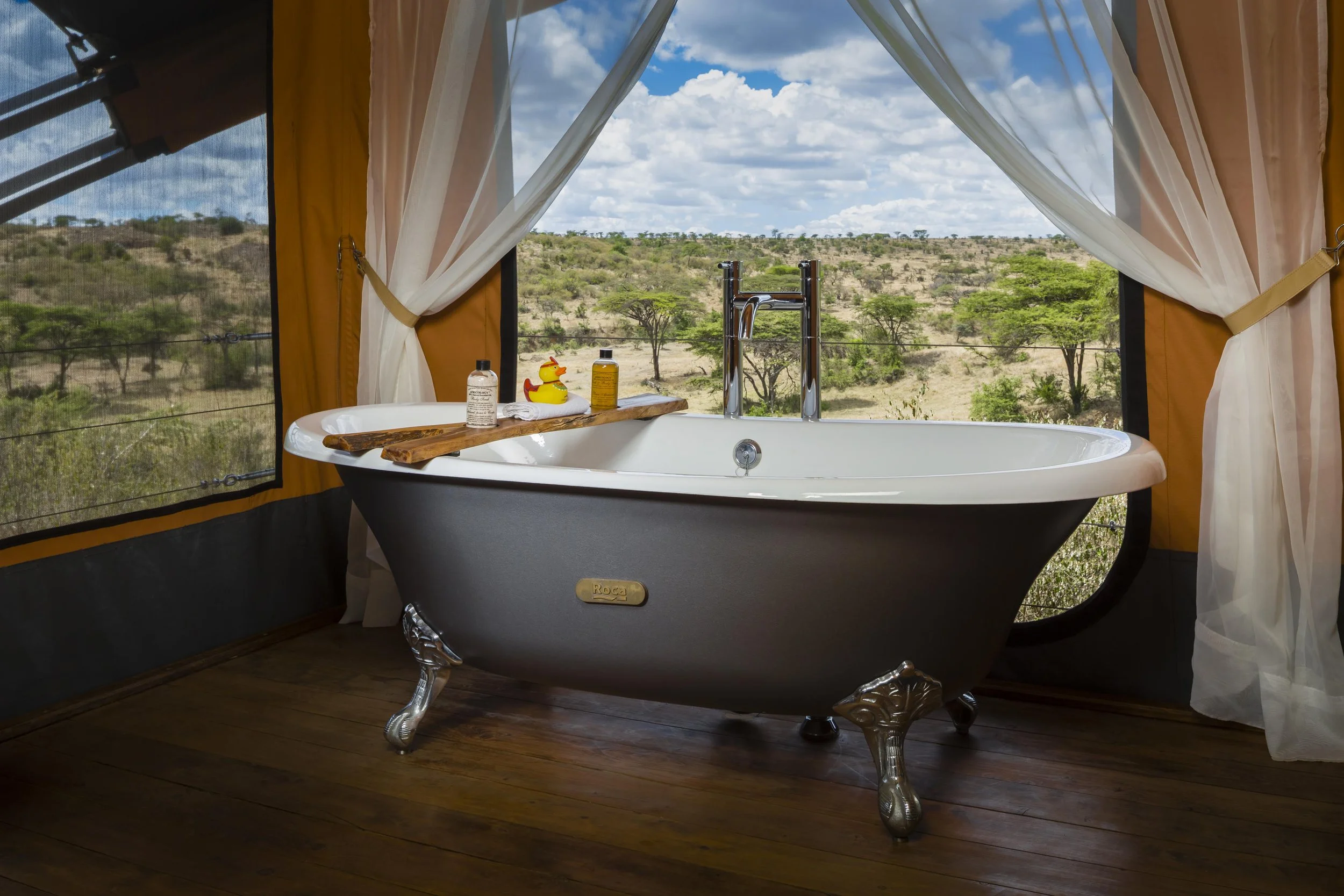 A black clawfoot bathtub with silver legs, placed inside a tent with large open window flaps and white curtains tied to the sides. There are some bath toiletries and a rubber duck on a wooden tray across the tub. Outside view shows a savannah landsca