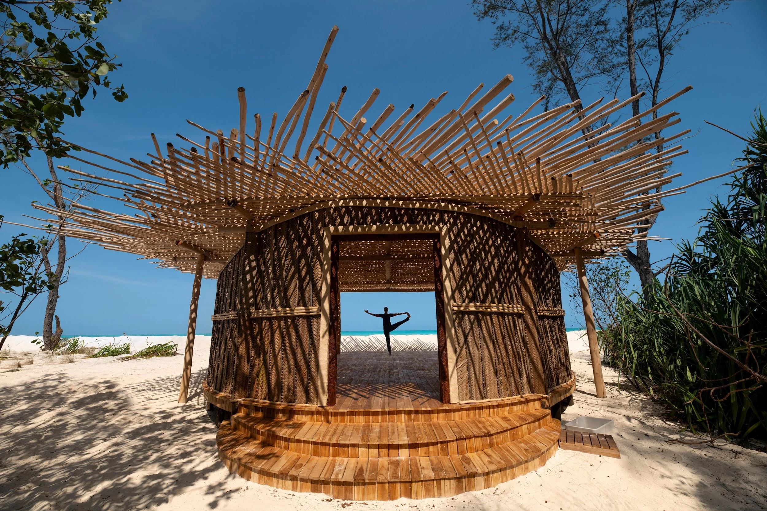 A circular hut made of woven wood and bamboo, with a large sunshade made of bamboo sticks radiating outward on a sandy beach with trees in the background, and a person practicing yoga inside.