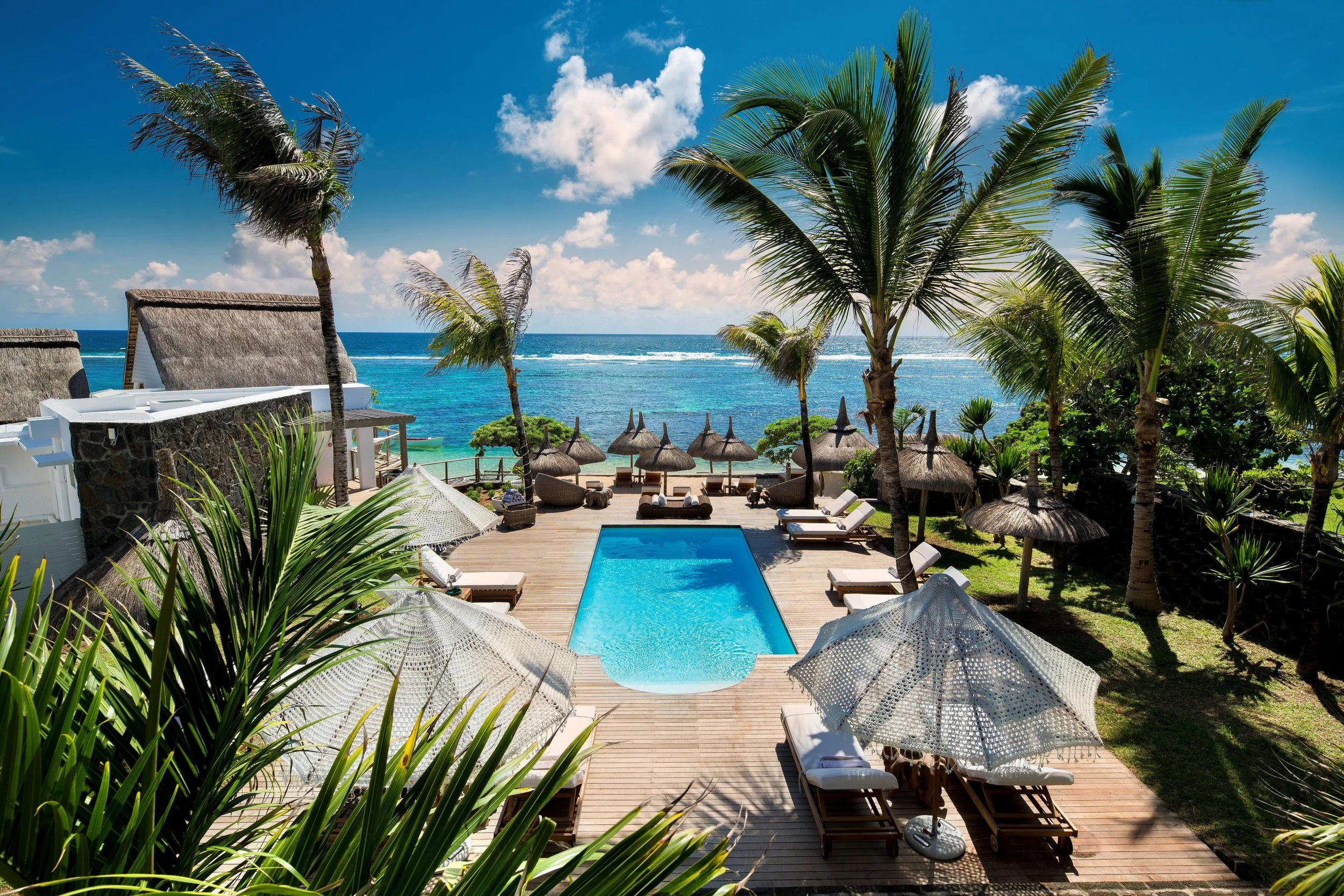 A tropical beach resort with a swimming pool, palm trees, lounge chairs with umbrellas, and thatched umbrellas overlooking the ocean under a bright blue sky.