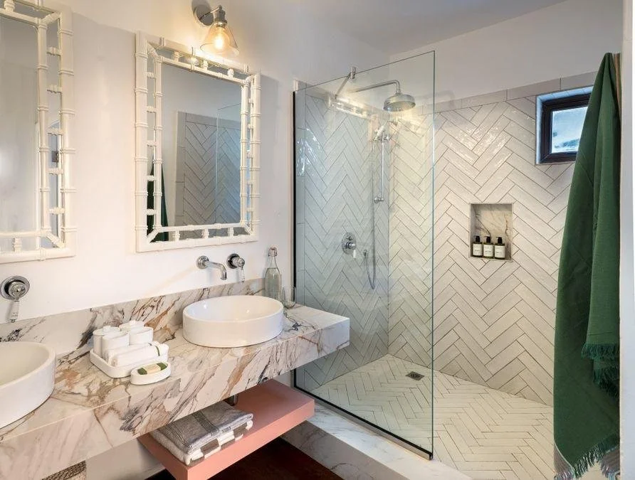 Modern bathroom with dual round vessel sinks on a marble countertop, framed mirrors, and a glass-enclosed shower with herringbone tile pattern and built-in niche for toiletries. A green towel hangs on a hook and small window is present near the ceili