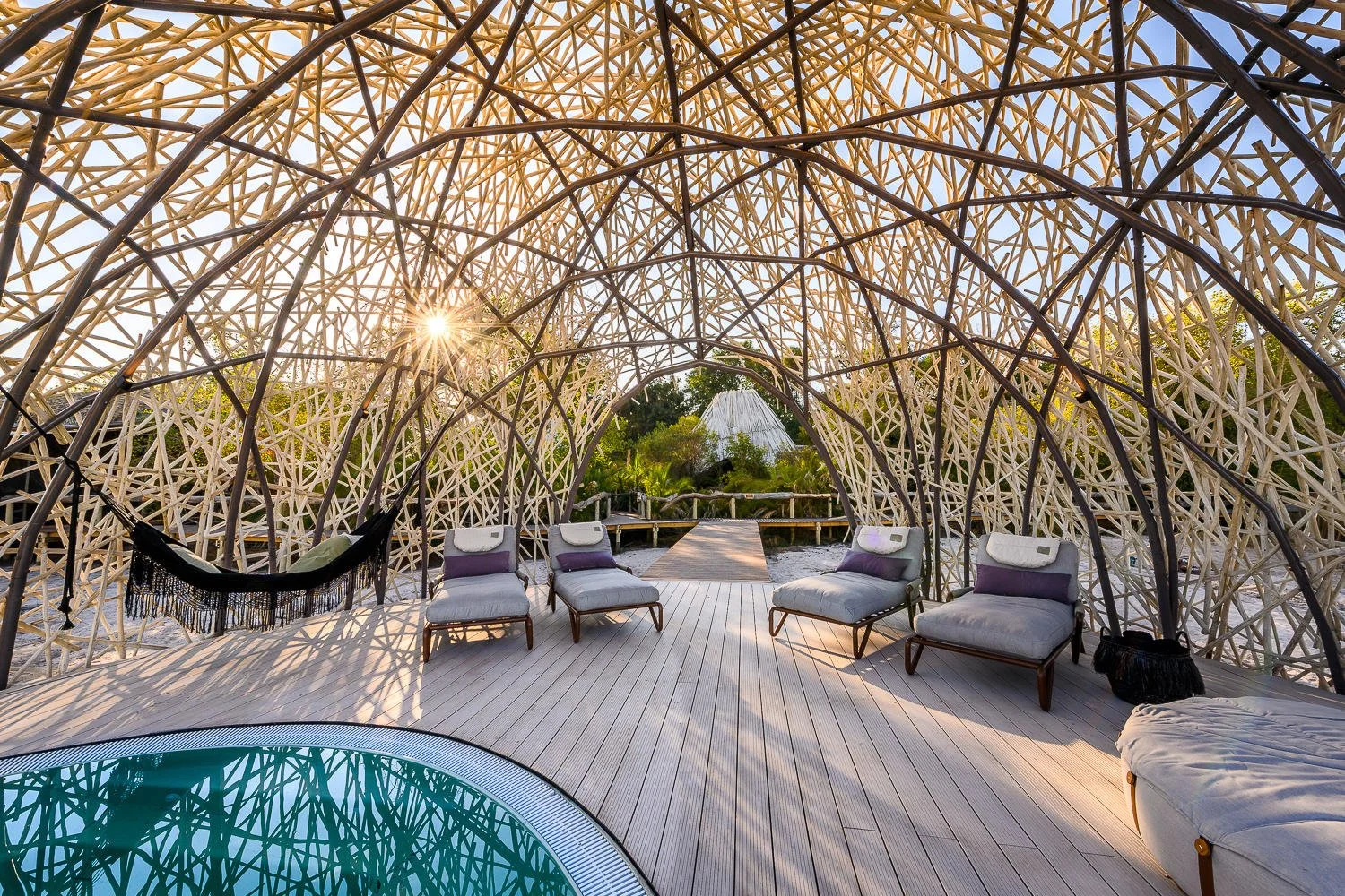 A relaxing outdoor space with a wicker dome-shaped structure, sun setting in the background, lounge chairs with pillows, a hammock, and a small pool with blue water.
