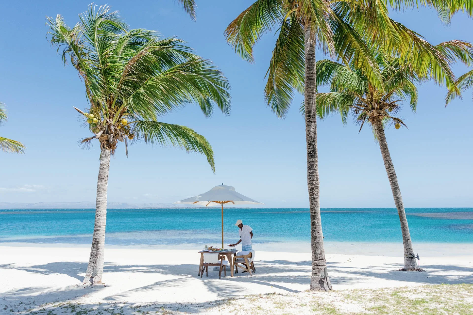 A tropical beach scene with white sand, palm trees, and clear blue ocean water; a person stands near a table under a large umbrella.