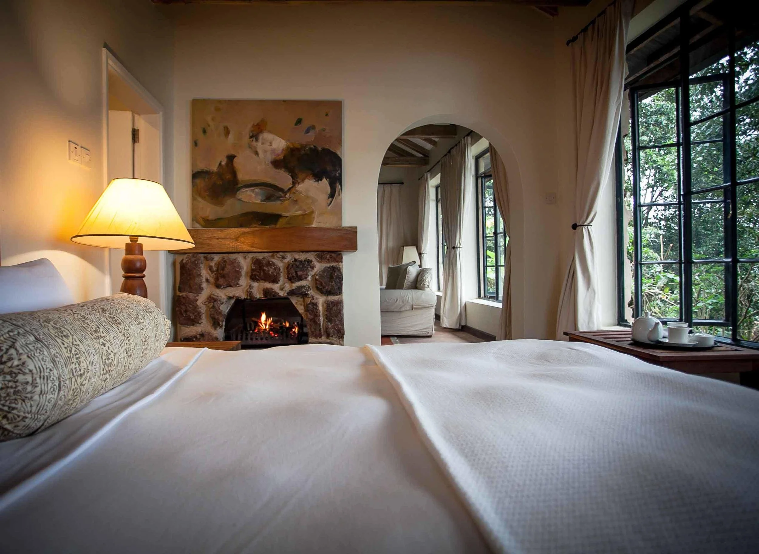 Cozy bedroom with a stone fireplace, bed with decorative pillow, lamp, and large windows with white curtains showing greenery outside.