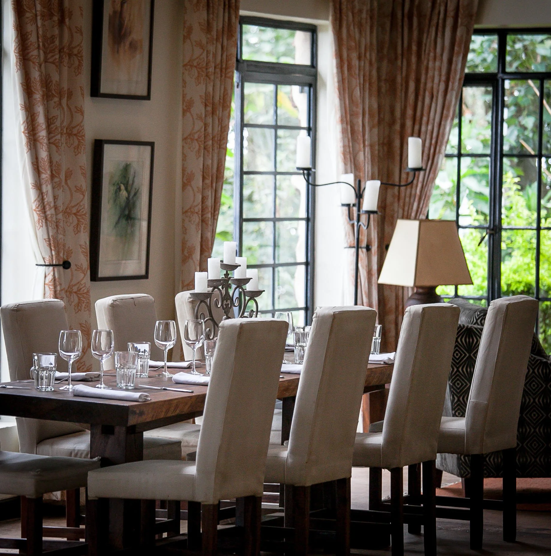 A dining room table set with wine glasses, water glasses, and napkins with a candelabra centerpiece, near large windows with curtains, framed art, and a lamp.