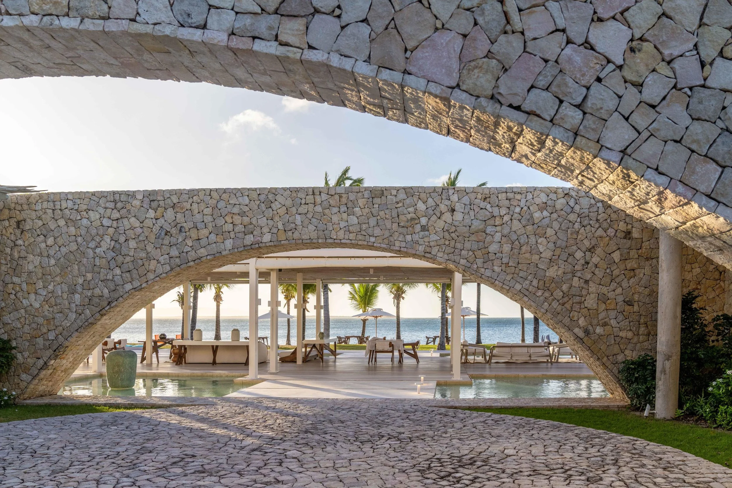 Stone arches framing a view of a pool area with lounge chairs, umbrellas, and palm trees overlooking the ocean at a beach resort.