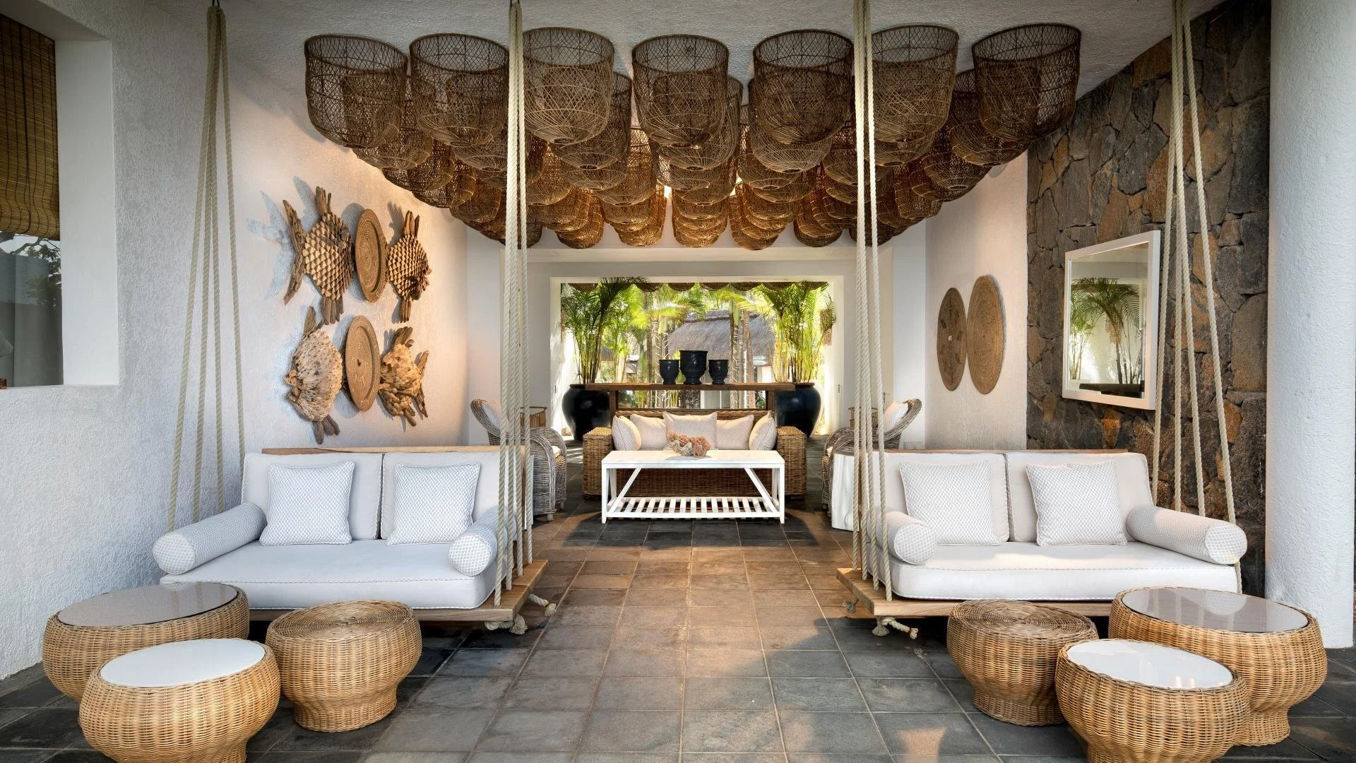 Tropical outdoor lounge area with white swing seats, rattan chairs, and tables, decorative coastal wall art, and lush greenery.