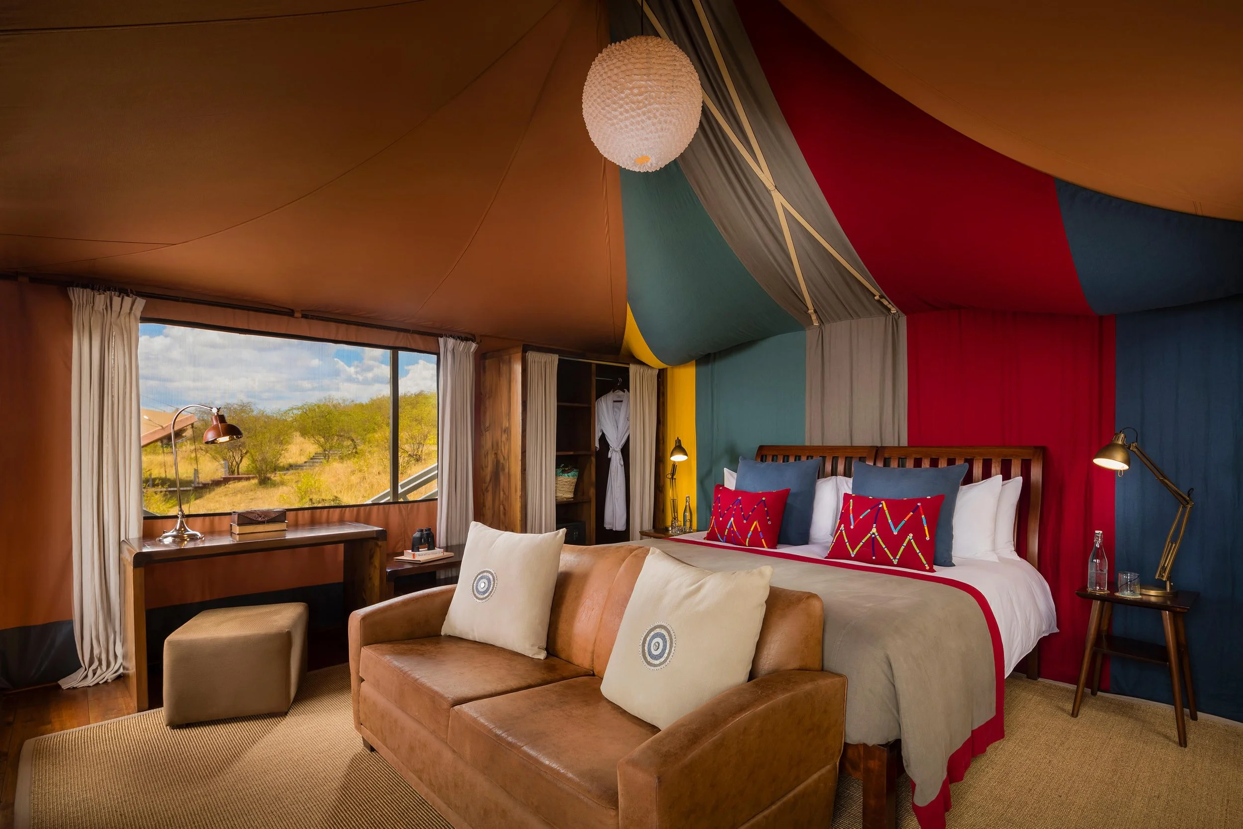 A bedroom with colorful fabric drapes on the ceiling and walls, a large window showing a scenic landscape, a bed with pillows, a tan leather sofa with cushions, a desk near the window, and warm lighting.