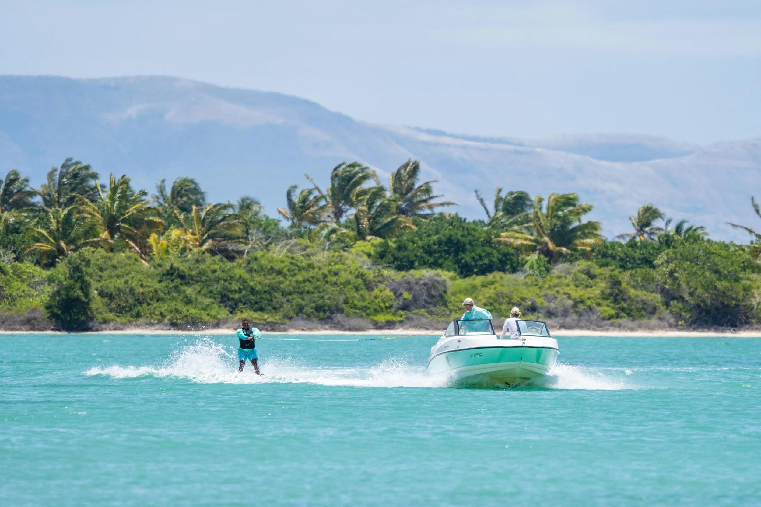 A motorboat speeding across turquoise water with two people onboard. A man waterskiing behind the boat, with a lush green island and mountains in the background.