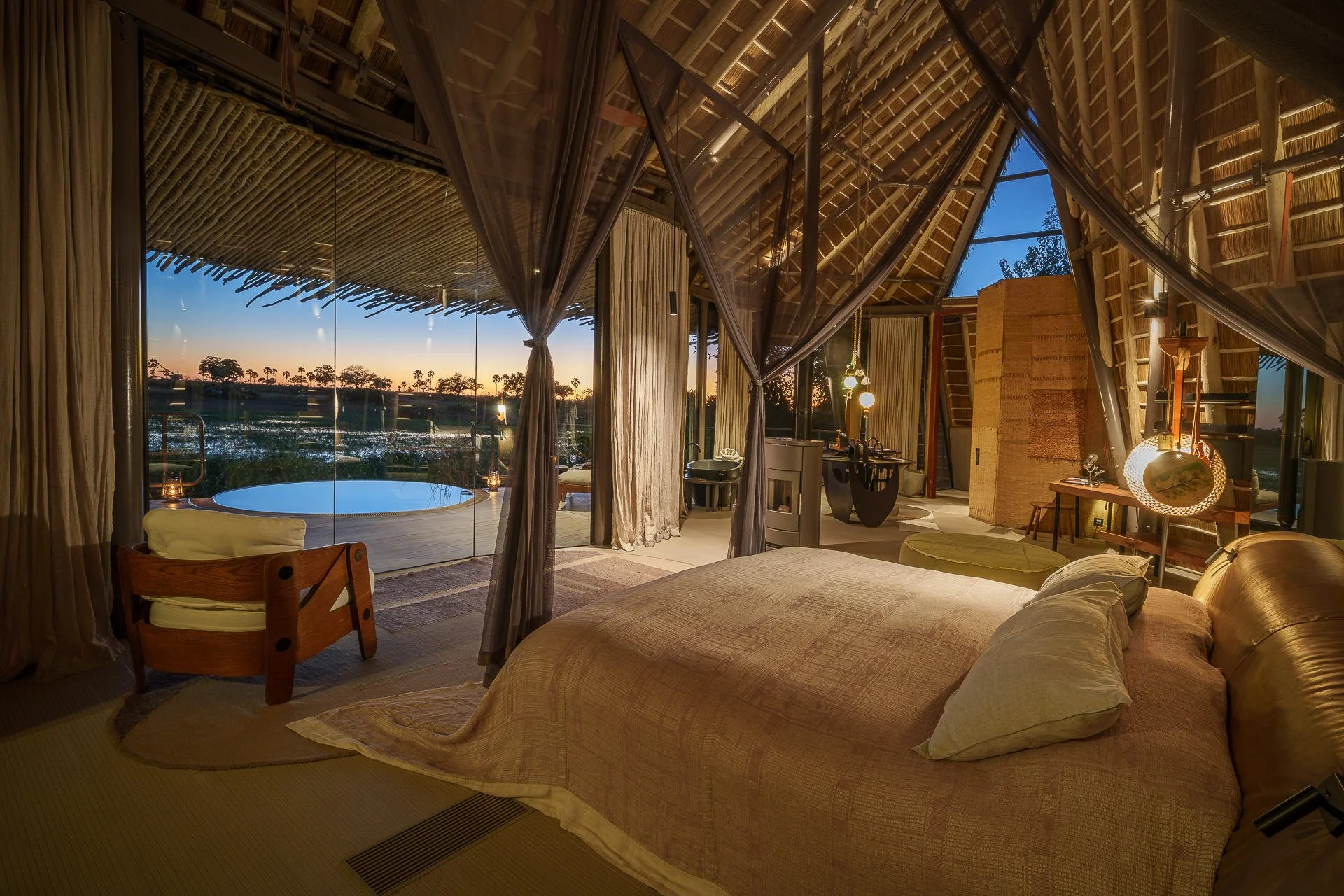 A luxurious bedroom with a large bed, curtains, and a backyard hot tub with a view of the sunset over a field and trees. The room has warm lighting, furniture, and decor with tropical design elements.