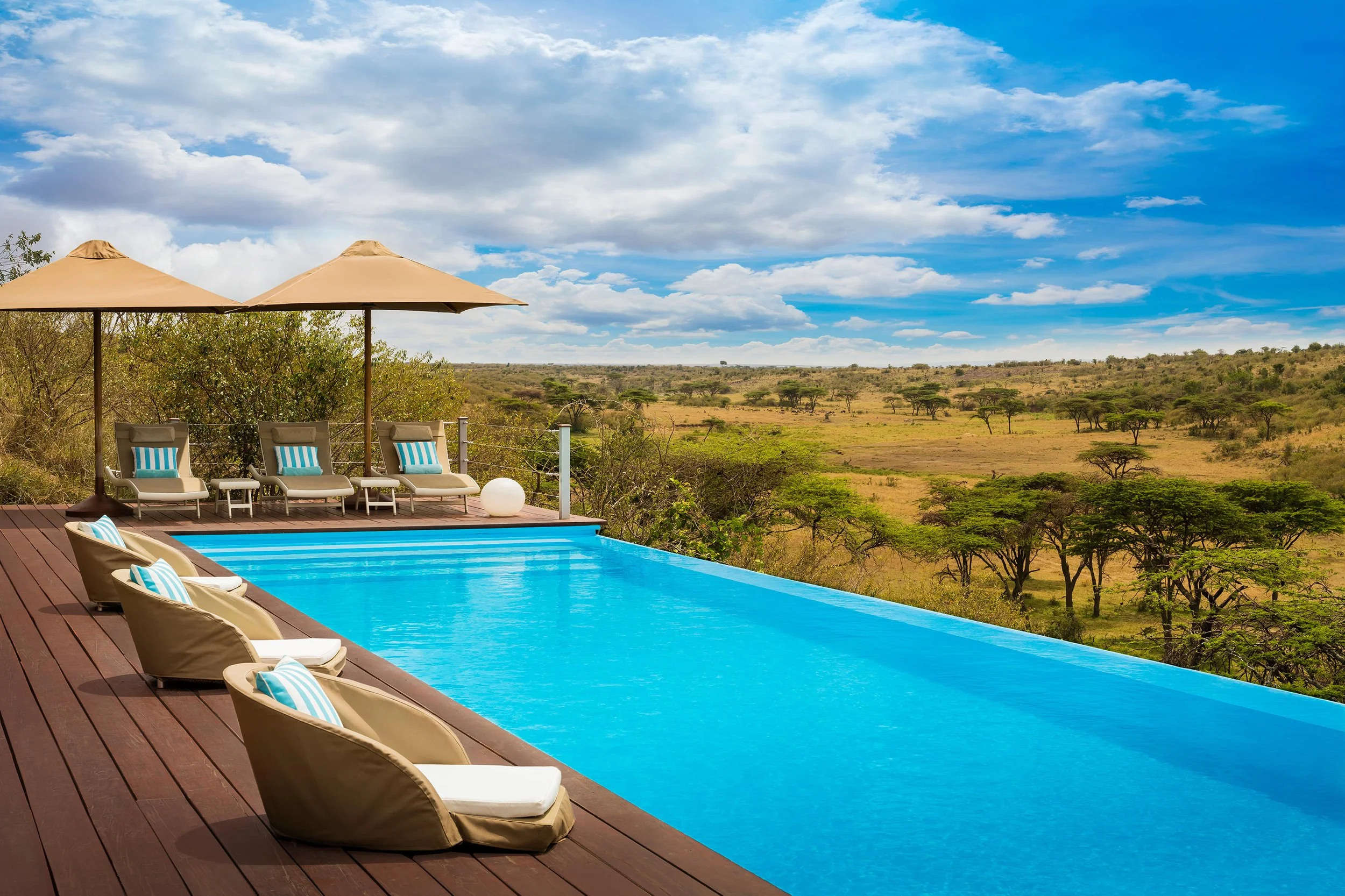 Luxury infinity pool on a wooden deck with sun loungers, umbrellas, and a view of African savannah with scattered trees and blue sky.