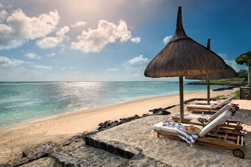 Beach scene with wooden lounge chairs, straw umbrellas, and a calm ocean under partly cloudy sky.