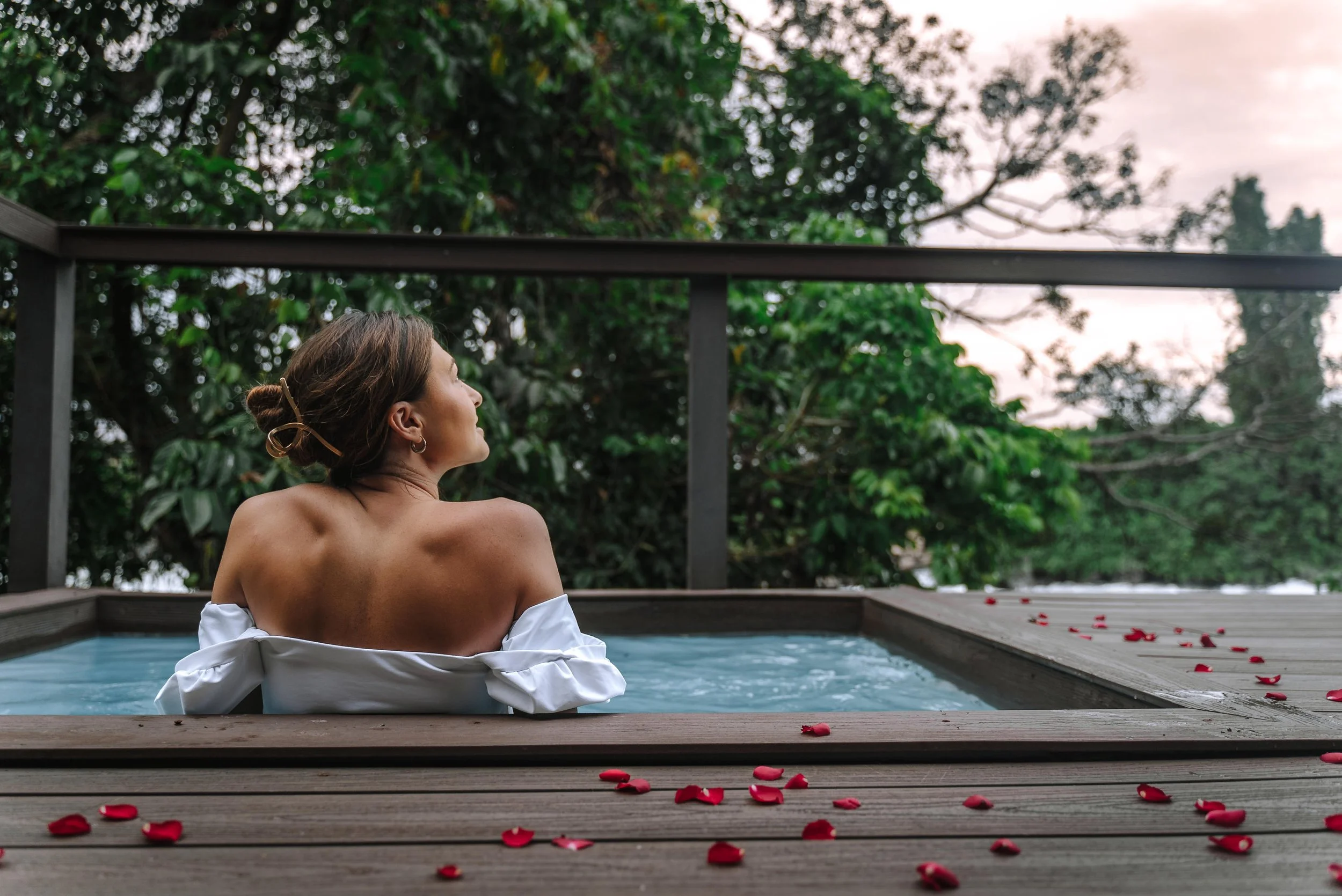 A woman with short brown hair styled in a bun is sitting in an outdoor hot tub, surrounded by rose petals, overlooking a green forest at dusk or dawn.