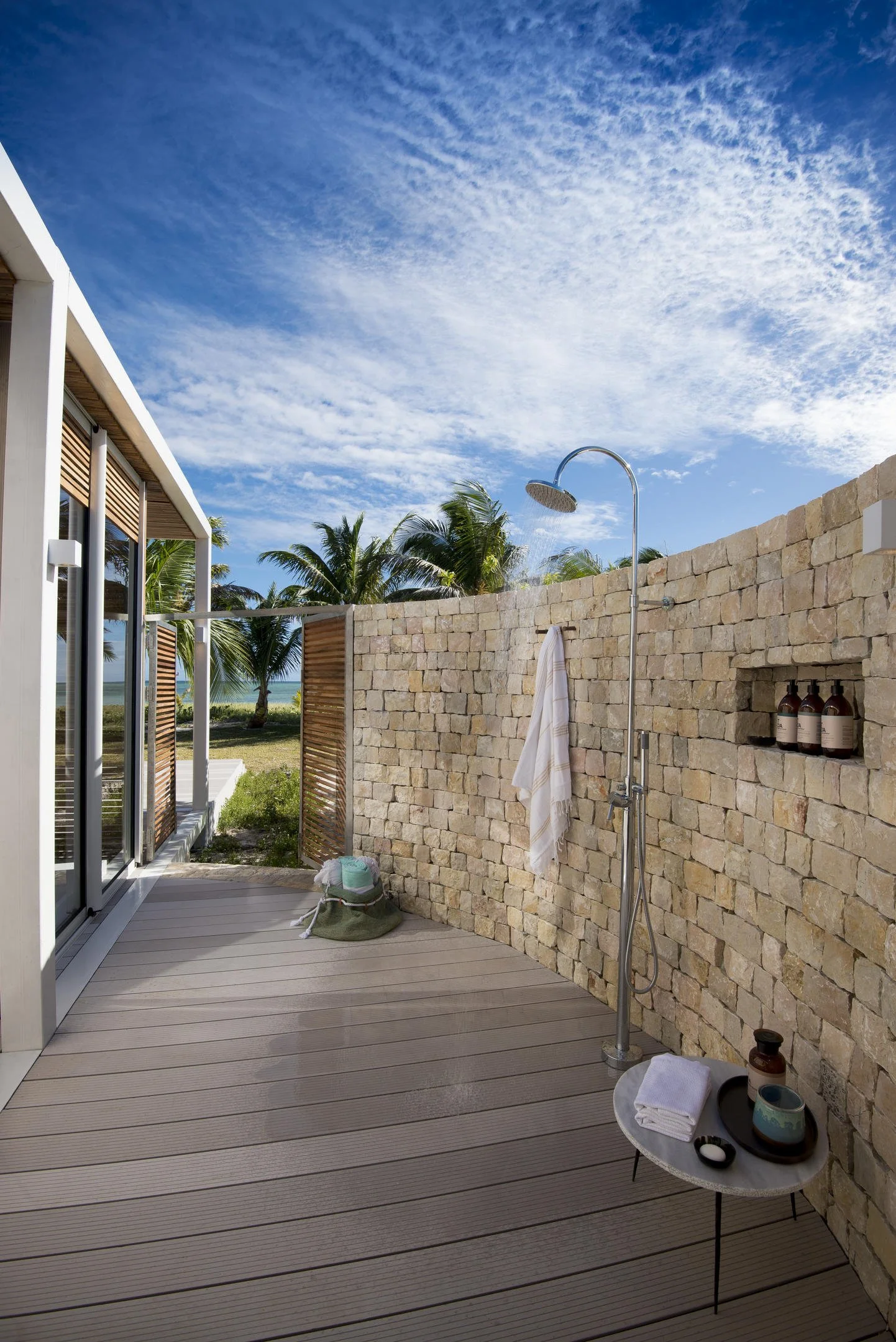 Outdoor shower area with a stone wall, a rain shower head, a towel hanging on the wall, and bottles on a built-in shelf, with trees and a blue sky in the background.