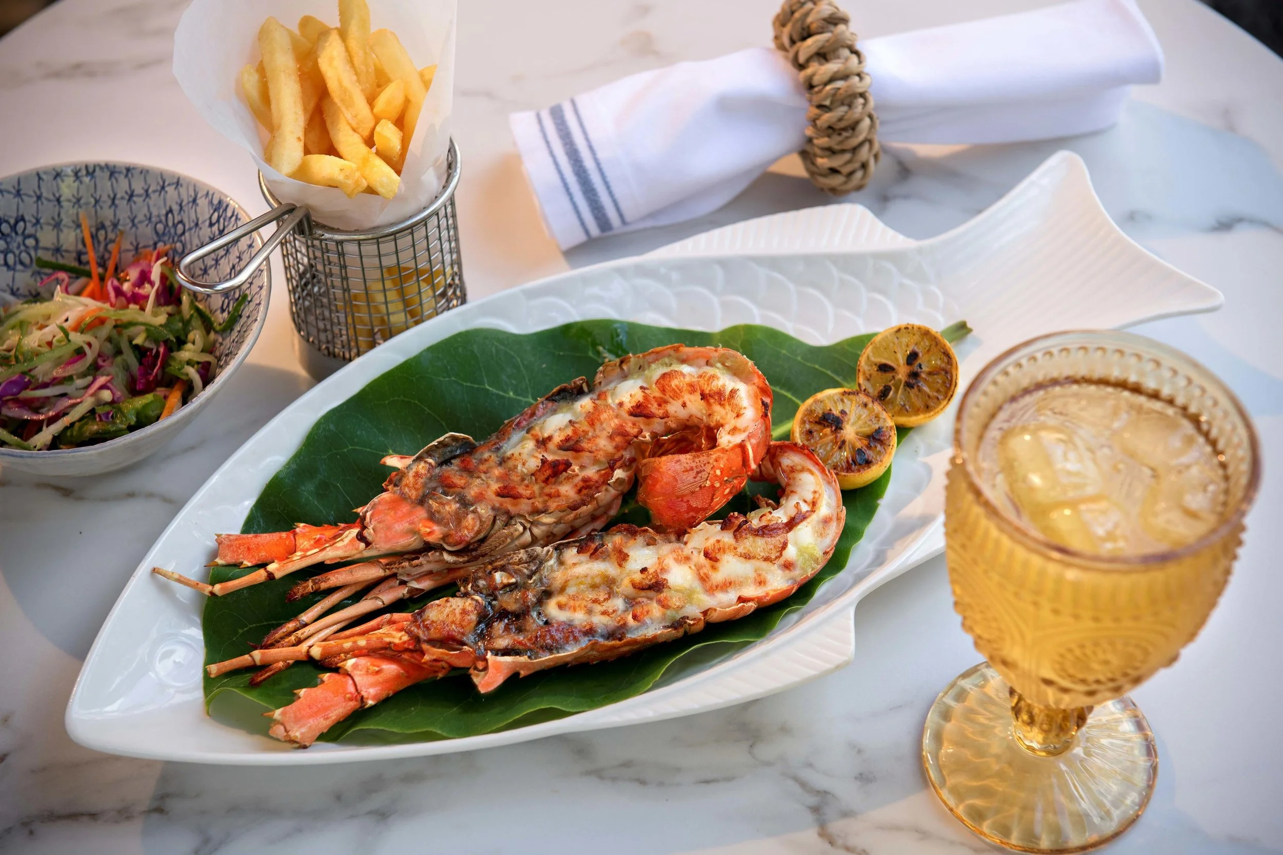Grilled lobster tails with grilled lemon slices on a white fish-shaped plate, served with a tomato-based drink in a vintage glass, alongside a side salad, French fries in a metal holder, and a folded napkin with a napkin ring on a white marble table.