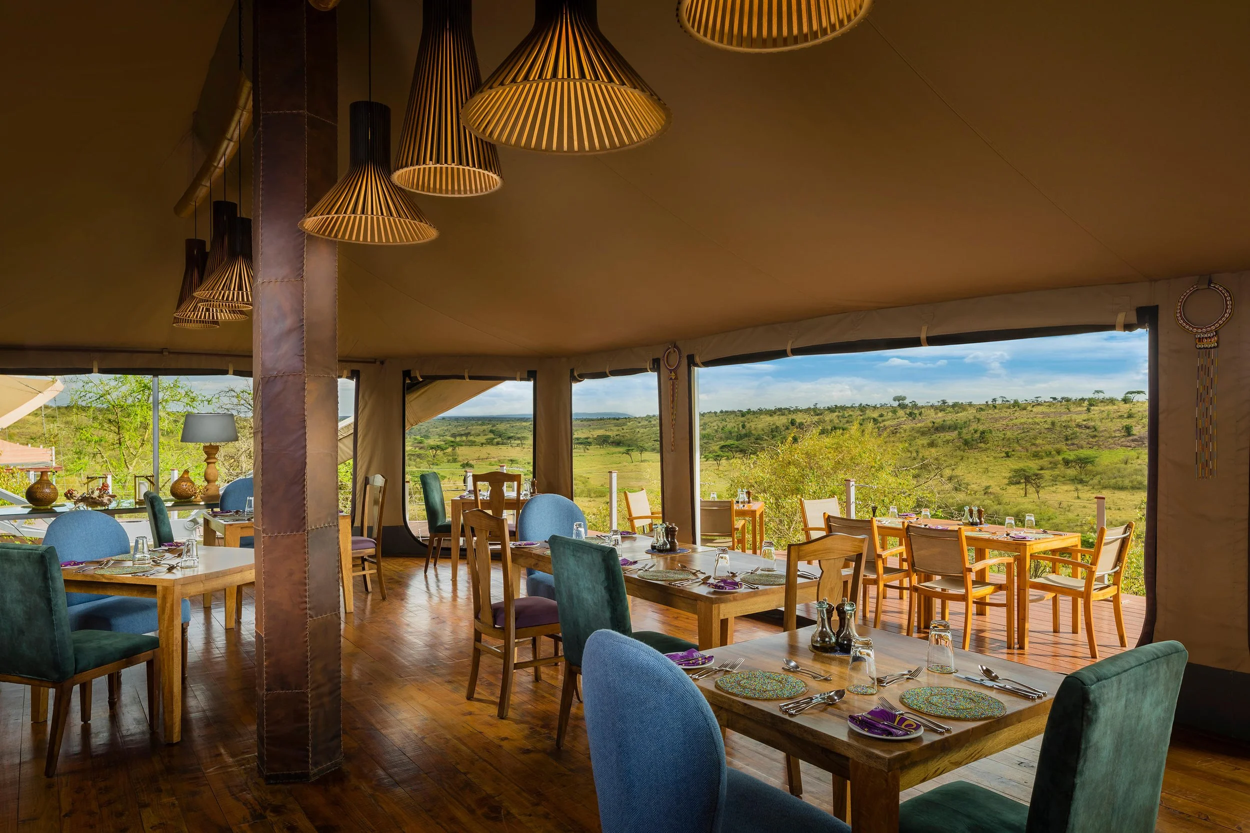 Interior of a restaurant with large windows showing a savannah landscape, wooden tables set with plates, glasses, and utensils, colorful chairs, and hanging pendant lights.