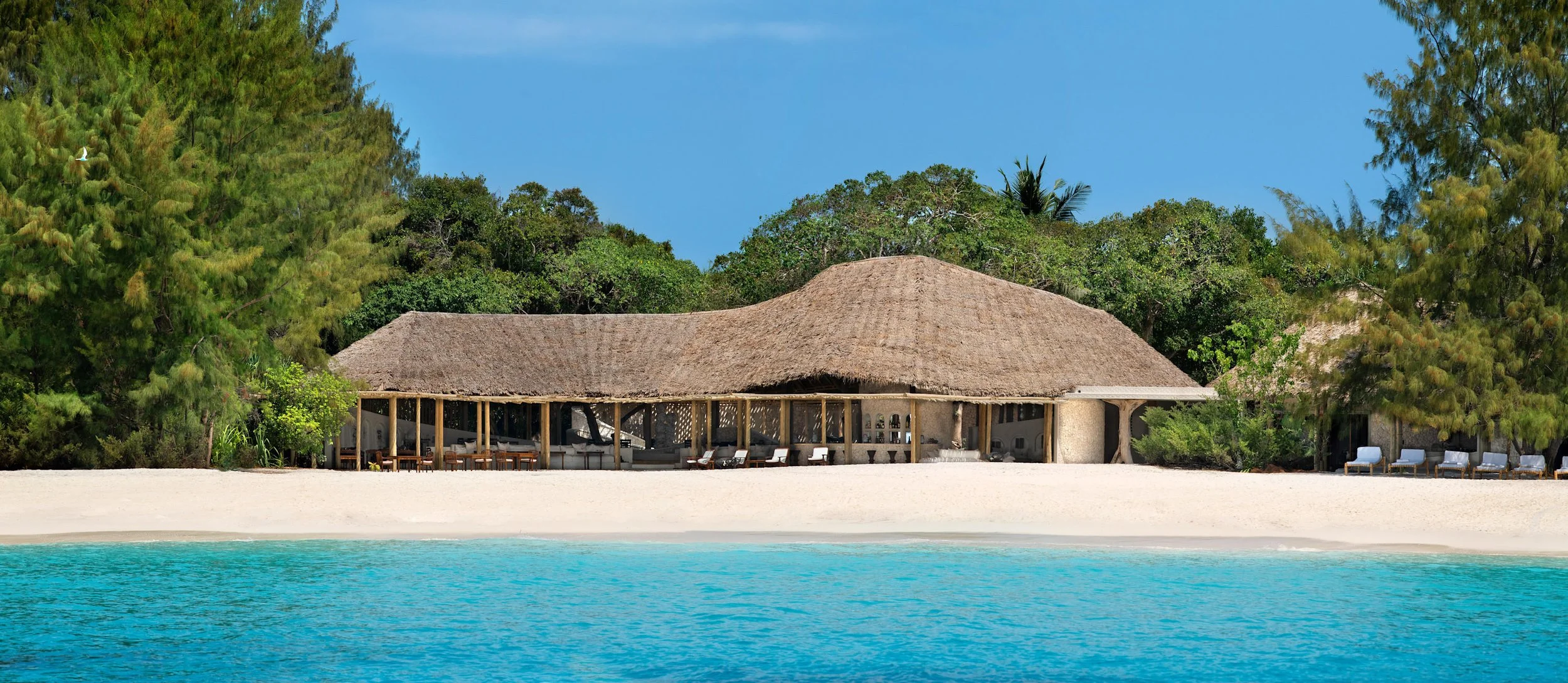 A tropical beach resort with a thatched-roof pavilion, white sand beach, and turquoise water, surrounded by lush green trees under a blue sky.