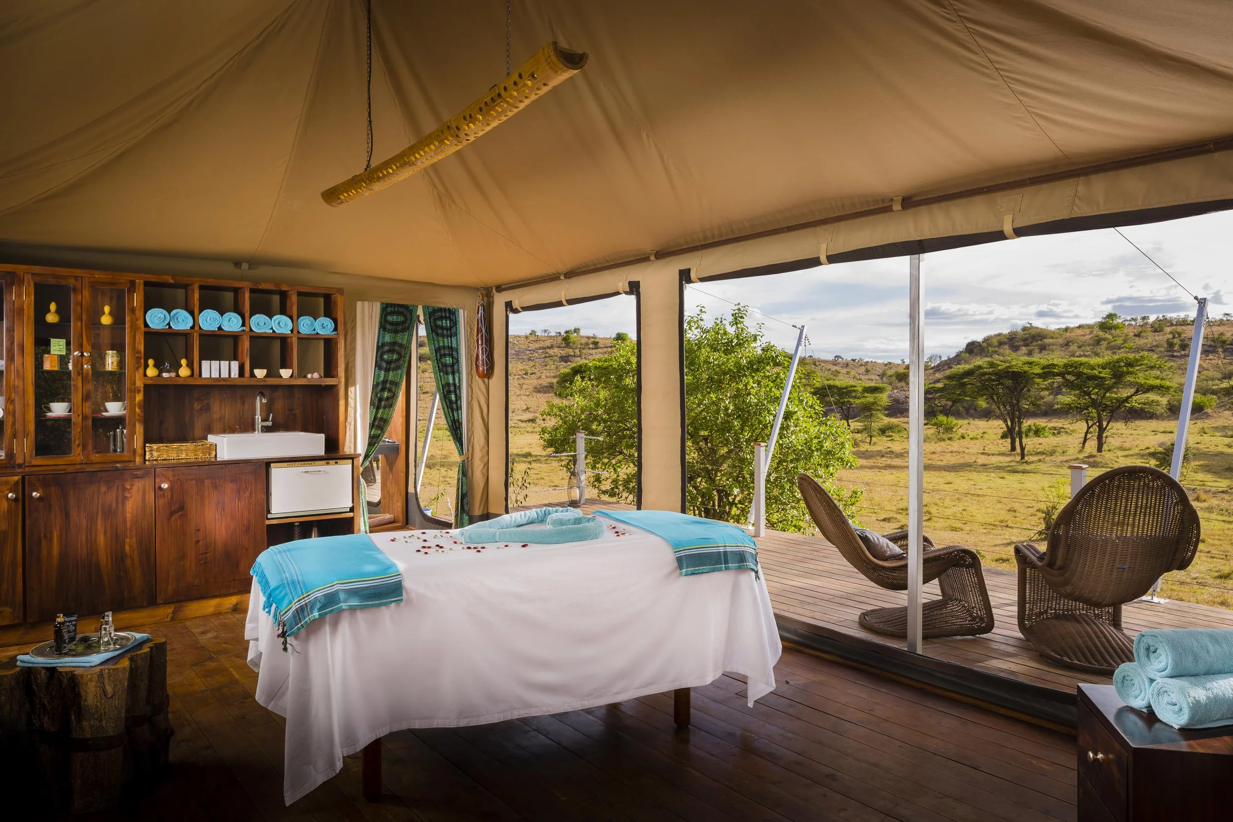 Luxury outdoor safari tent with bed, wooden furniture, seating area, and scenic view of savannah landscape with trees.