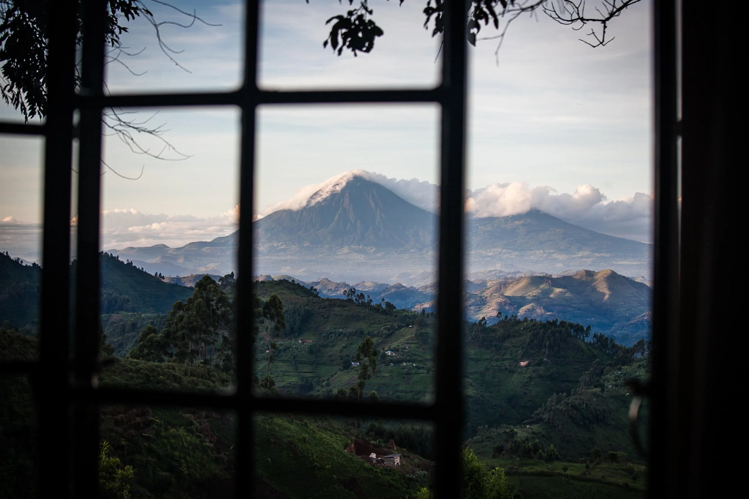 Mountain landscape viewed through a window with metal bars, showing a snow-capped volcano surrounded by green hills and scattered clouds.