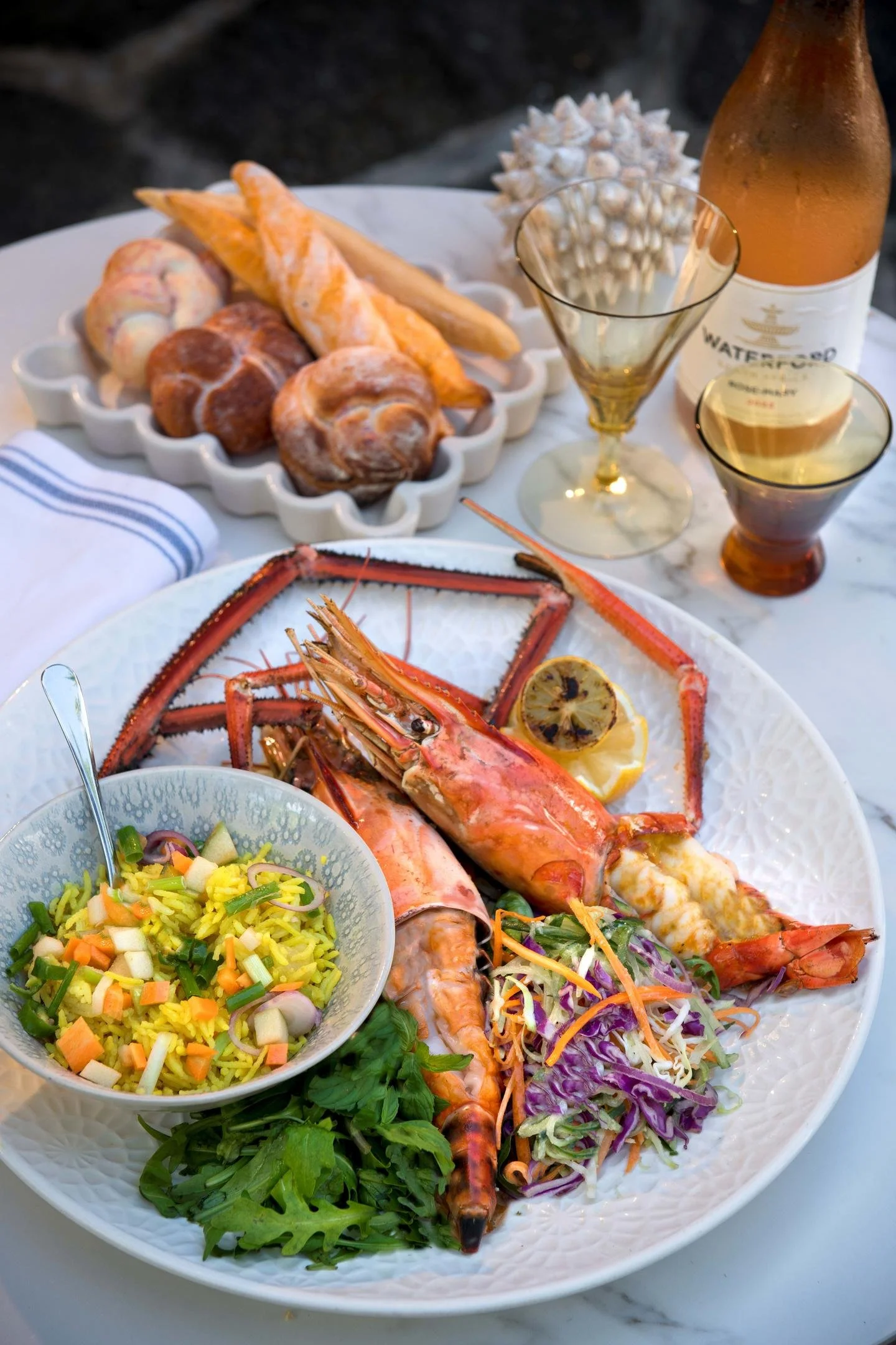A seafood feast featuring a large plate of lobster, shrimp, and salad, with lemon slices, and a side of bread rolls with butter, accompanied by a bottle of rosé wine and two glasses on a white marble table.