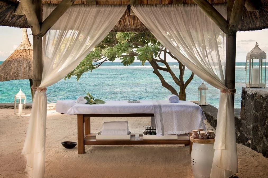 Beachside outdoor spa setup with a massage table under a canopy, surrounded by lanterns, decorative jars, and a view of the ocean and a tree.